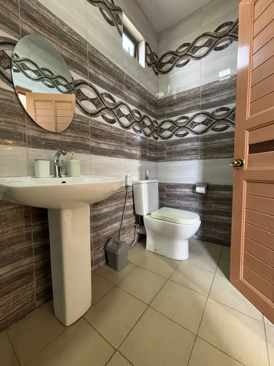 A small bathroom with a corner glass shower enclosure, white pedestal sink with a green hand soap dispenser, a small mirror, a towel hanging on a rack, and a toilet. The walls have horizontal striped tiles in shades of gray and brown, and the floor has multicolored pebble-patterned tiles.