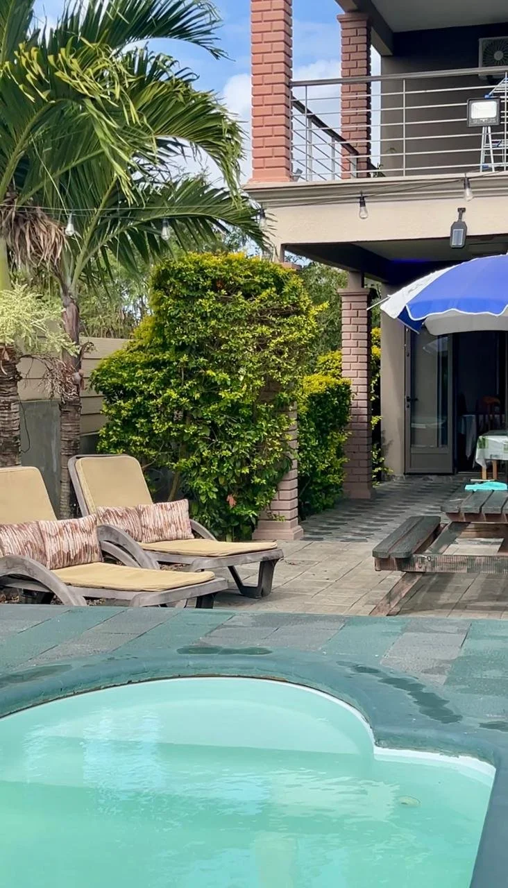 A backyard outdoor space with a hot tub in the foreground, beige lounge chairs with cushions, lush green bushes and palm trees, an awning with an umbrella, and a two-story house with brick columns and balcony.