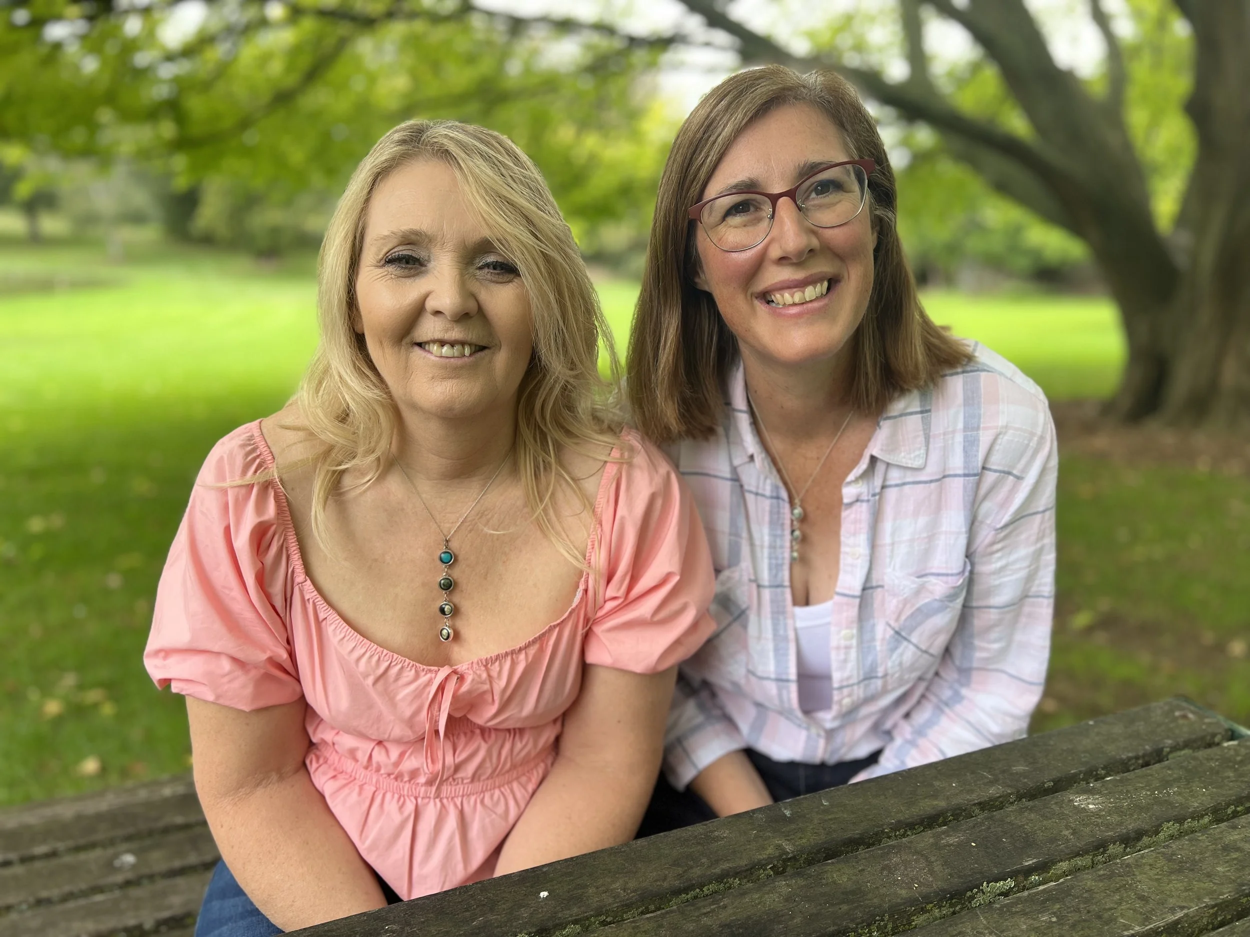 Two women sitting at a wooden picnic table outdoors in a park with green grass and trees, smiling at the camera.