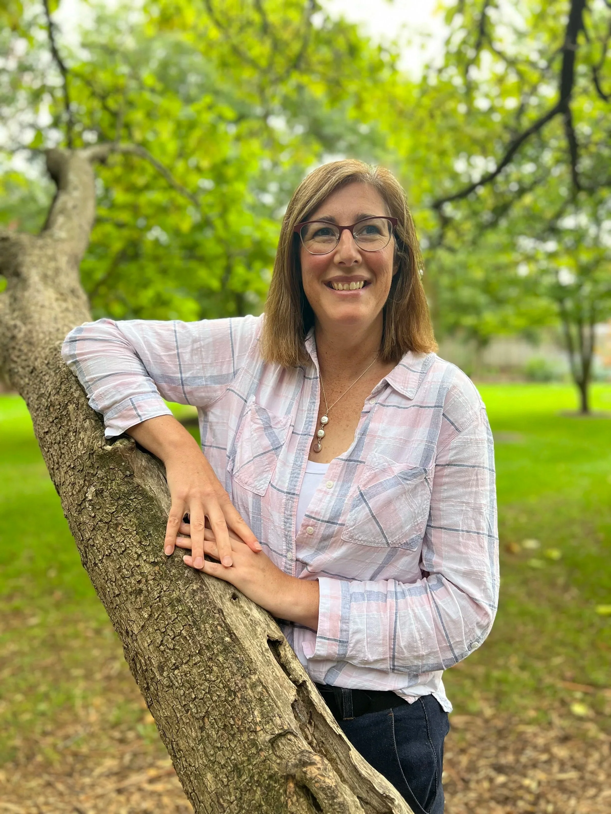 A woman with glasses and shoulder-length brown hair smiling outdoors, leaning on a tree with green leaves in a park.