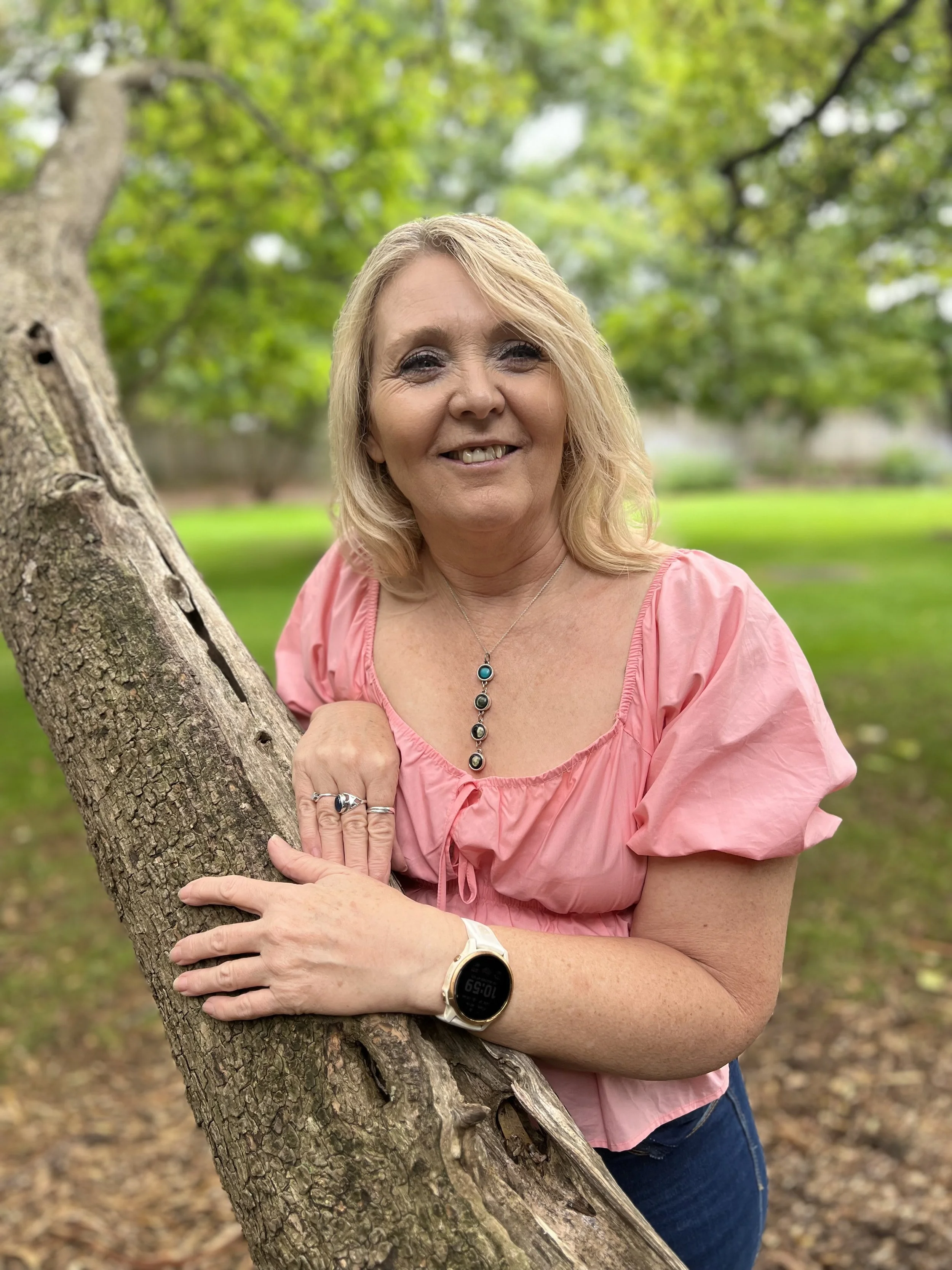 A woman with blonde hair smiling outdoors, leaning on a tree trunk in a park with green grass and trees in the background, wearing a pink blouse, jewelry, and a smartwatch.