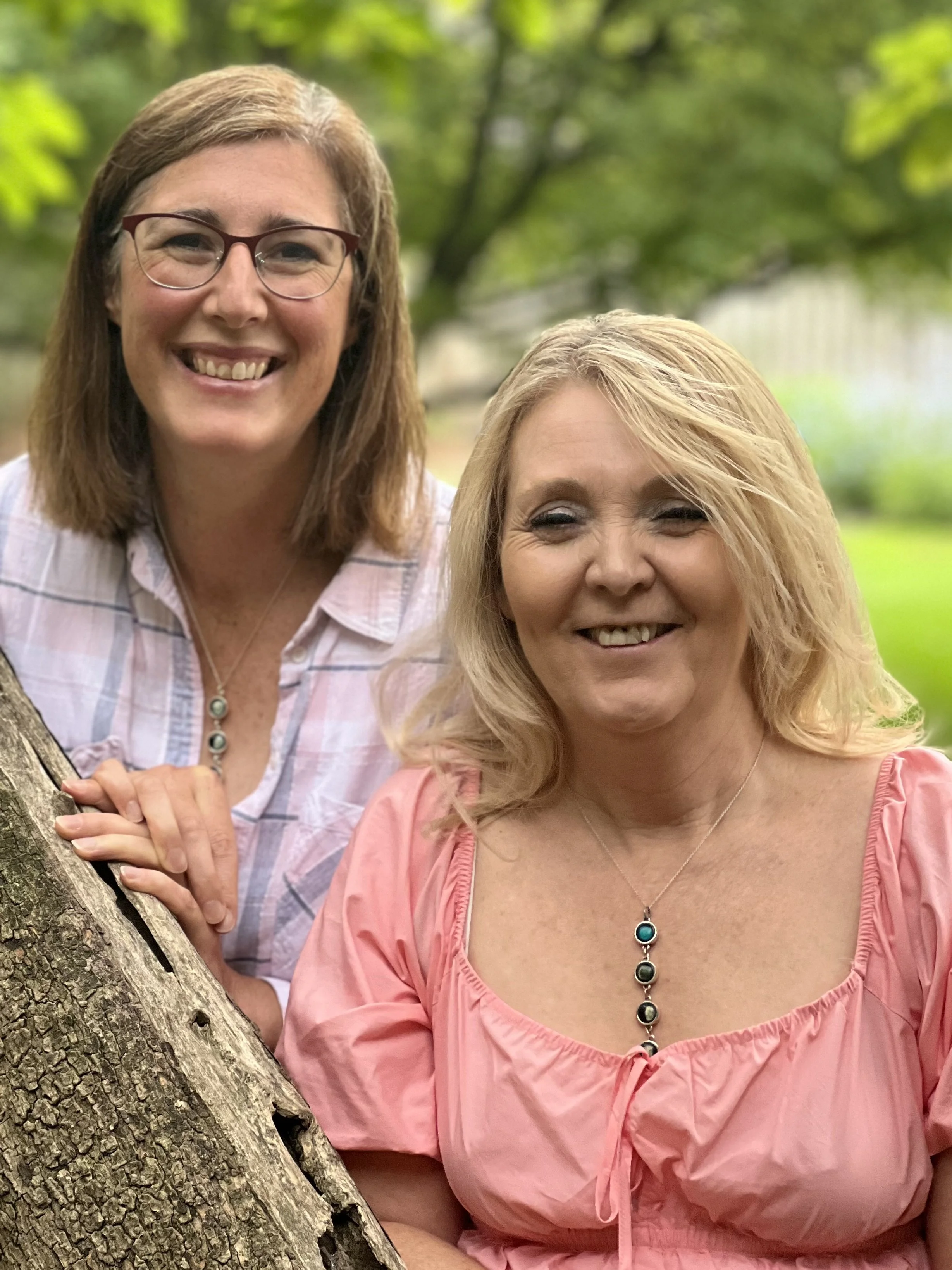 Two women outdoors, smiling and posing for a photo, with green trees and grass in the background.