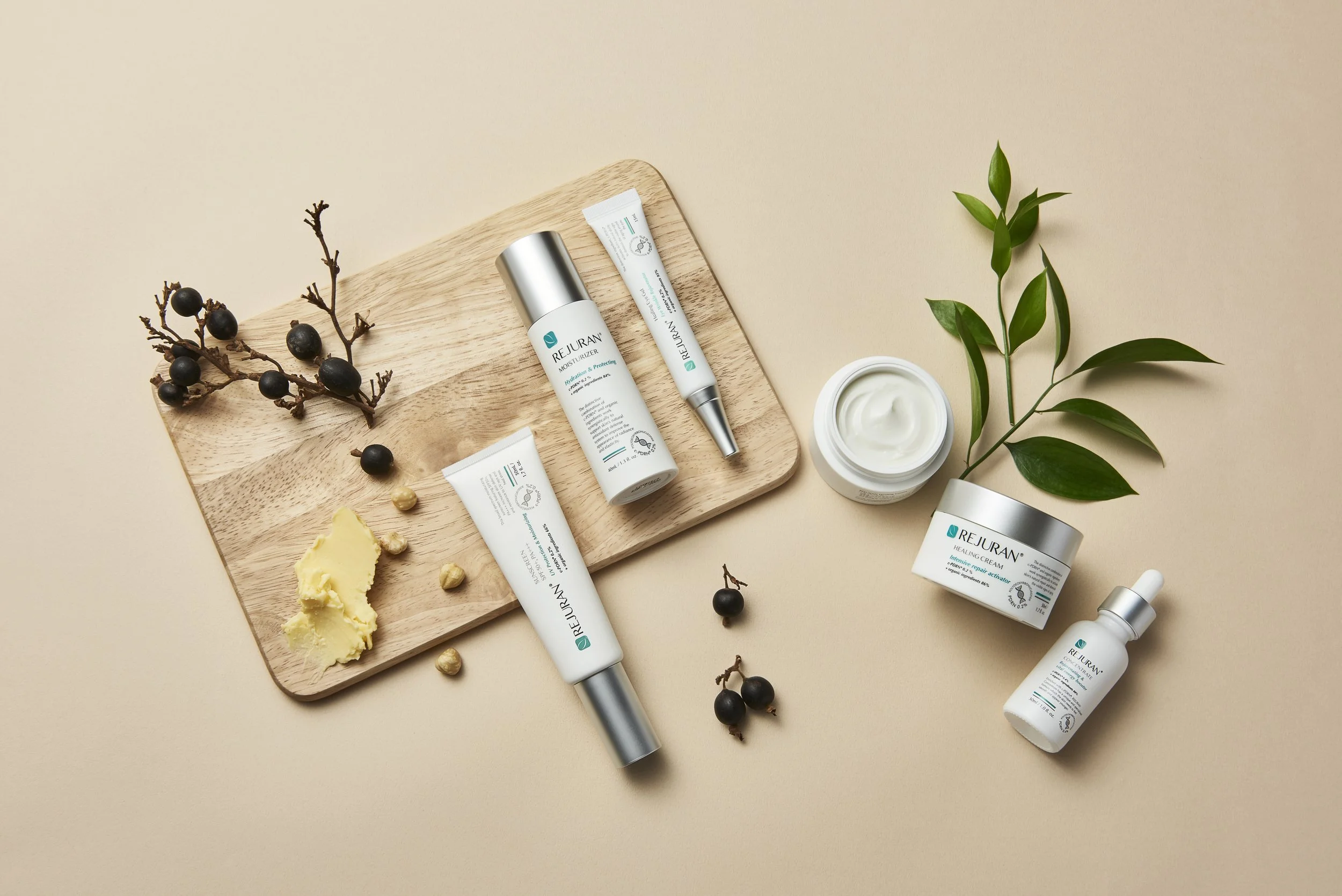 Assorted skincare products on a wooden tray, with a sprig of green leaves, black berries, pale yellow wax, and some small nuts, set against a neutral background.