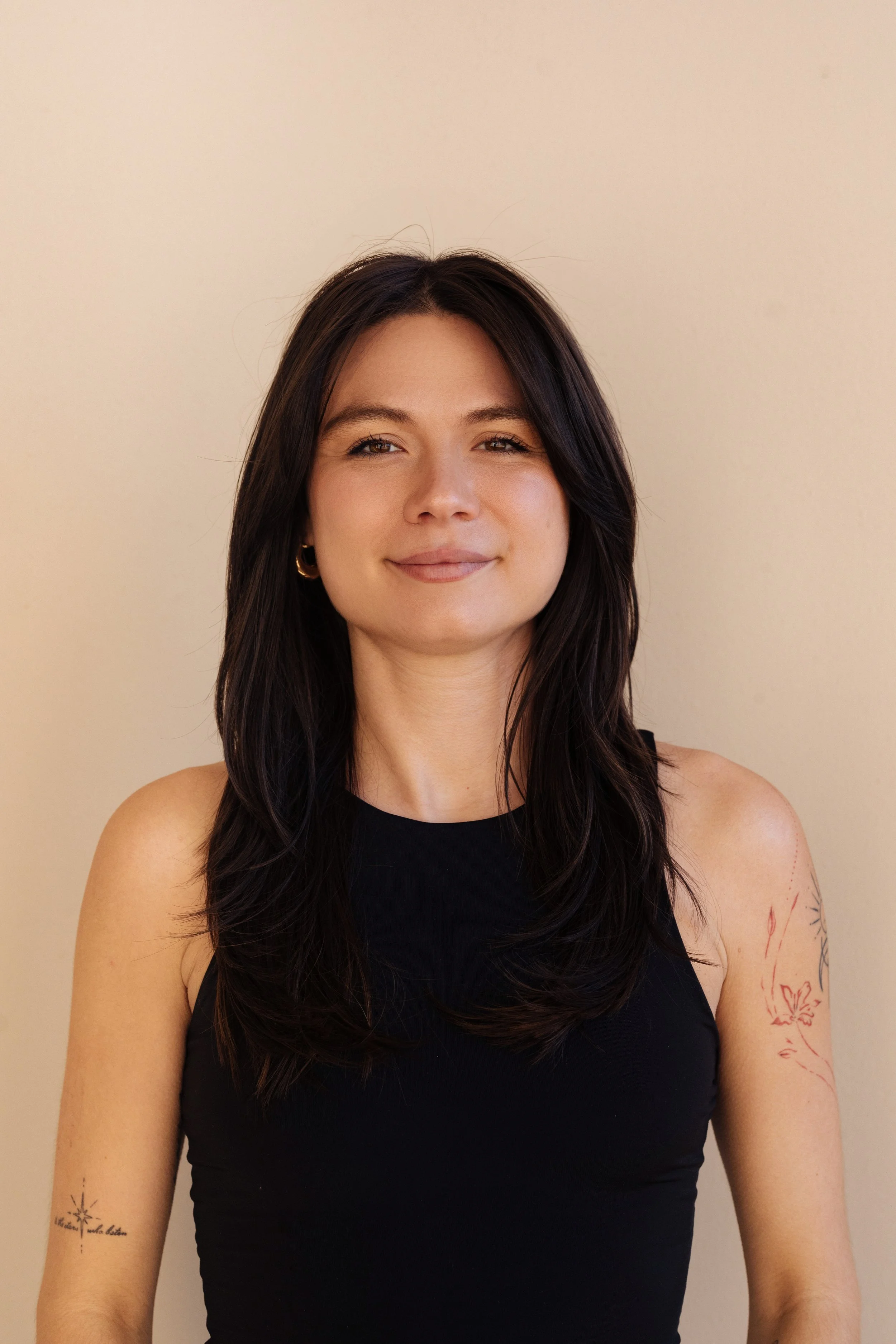 A woman with dark brown hair, wearing a black sleeveless top, standing against a plain beige background.