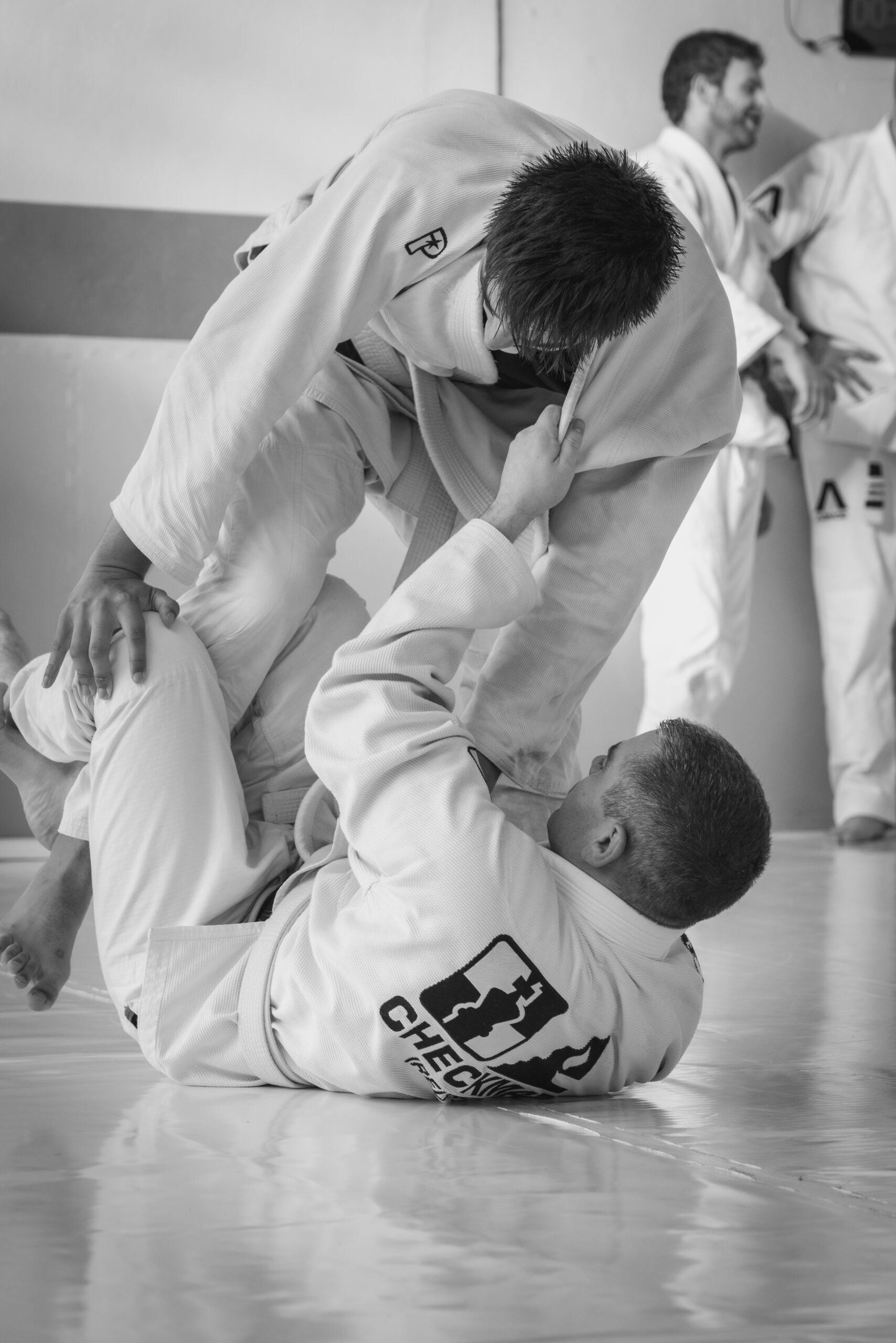 Two Brazilian Jiu-Jitsu practitioners in gis demonstrate a grappling technique on the mat, with other practitioners observing in the background.