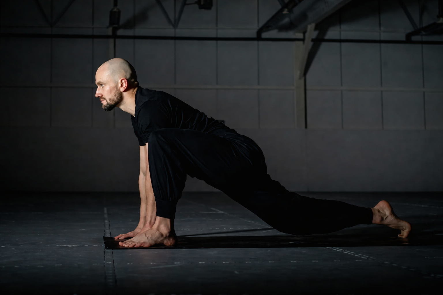 A man in black athletic clothing is on a black yoga mat, stretching in a spacious industrial-style room with gray walls and exposed pipes.