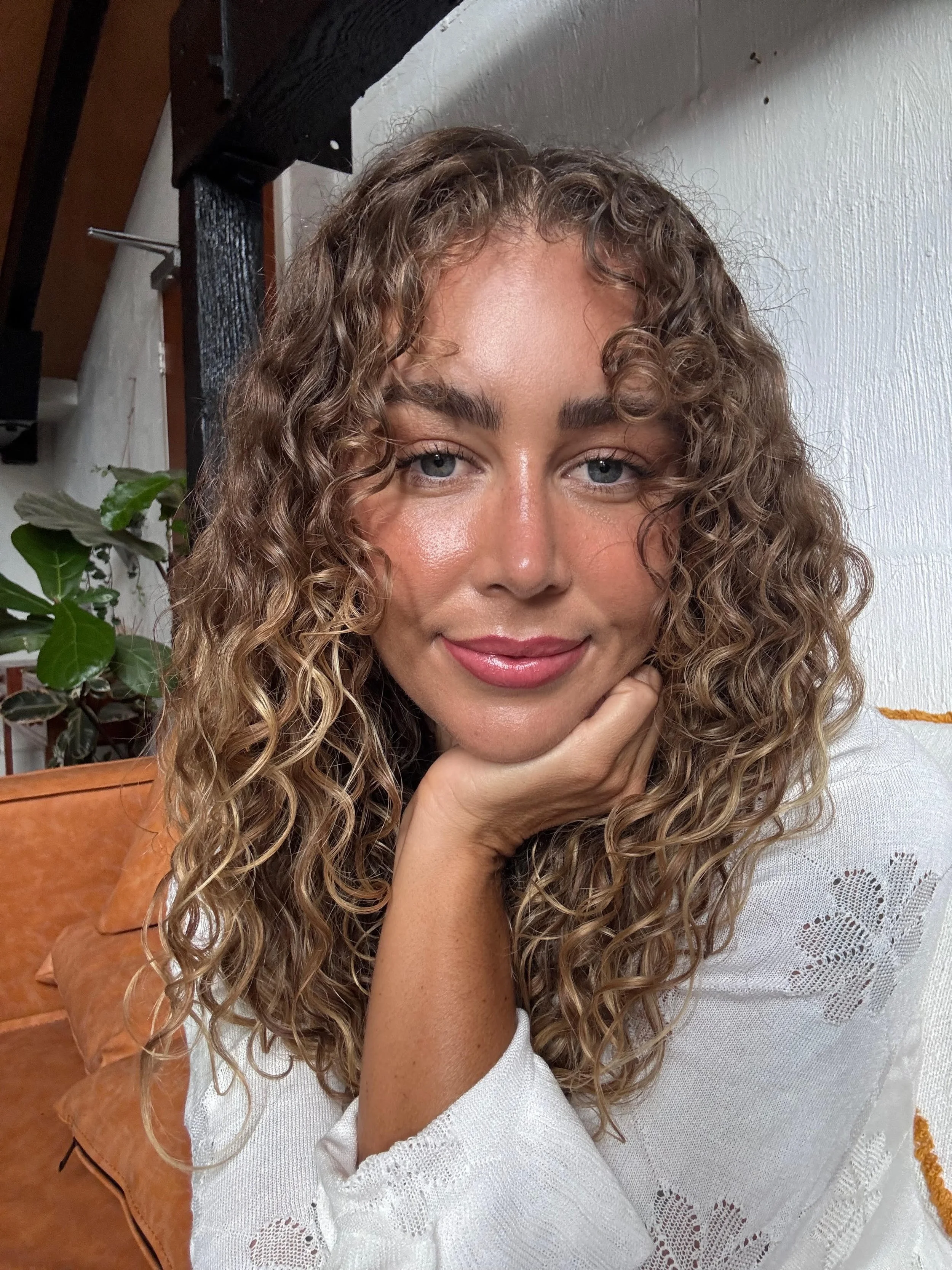 A woman with curly blonde hair, blue eyes, and light makeup sitting indoors with her chin resting on her hand, wearing a white top with lace details.