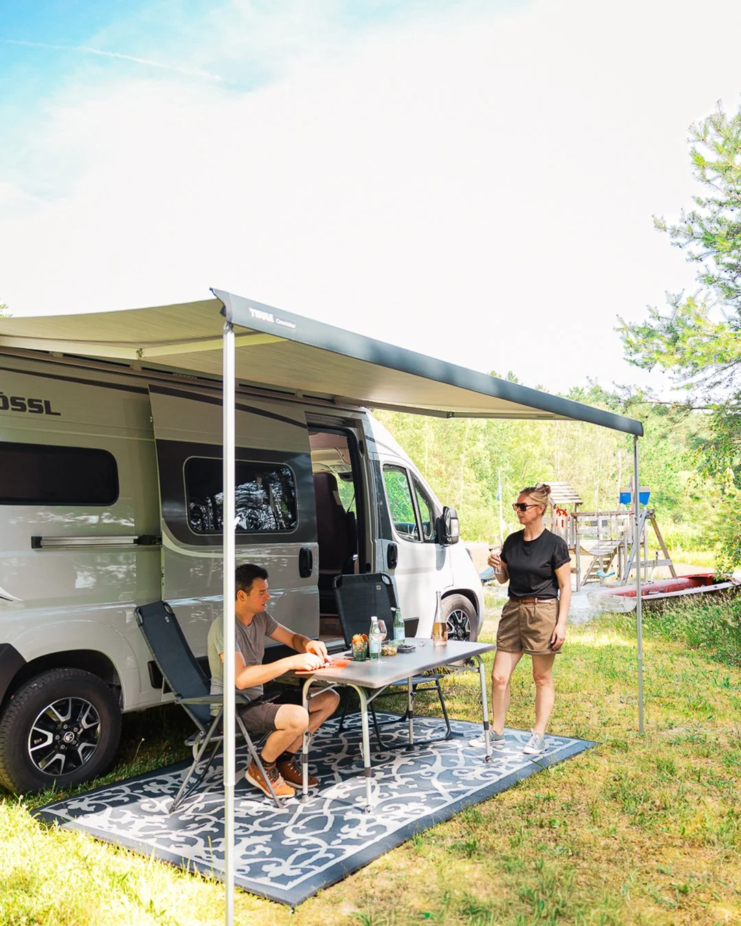 Mensen zitten en staan buiten een camper op een aangegeven plek in een groene omgeving, onder een uitschuifbare luifel, met tafel en stoelen, in een natuurlijke campingomgeving.