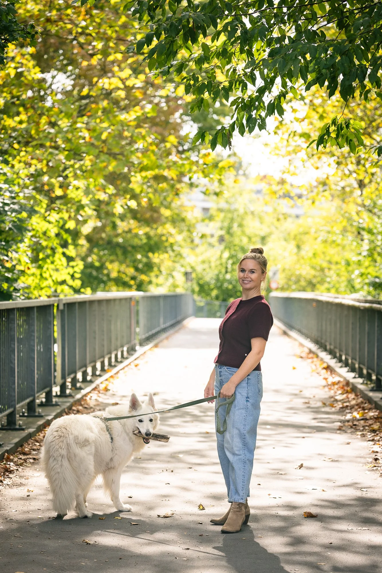 Vrouw wandelt met haar witte hond op een rustige brug omgeven door groene bomen in de herfst.