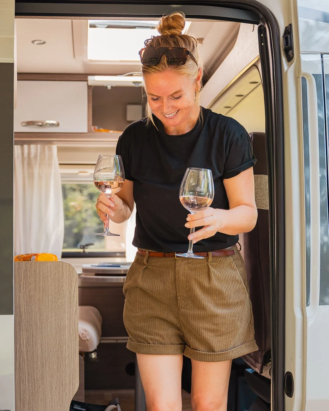 Vrouw met een topknot en zonnebril op haar hoofd die inside een camper rosé wijn drinkt.