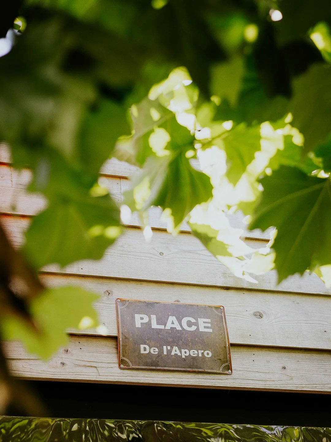Een houten muur met een metalen plaatje waarop staat 'PLACE De l'Apero', zichtbaar tussen groene bladeren.