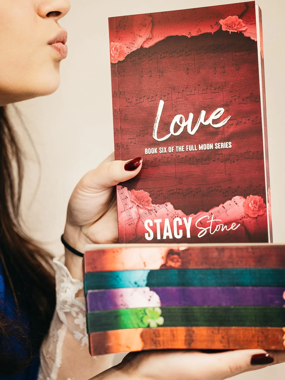 Een persoon houdt een boek met de titel 'Love' van Stacy Stone, boek zes uit de Full Moon-serie, met een rode cover en muzikale notities op de achtergrond, terwijl op de achtergrond andere kleurrijke boeken staan.