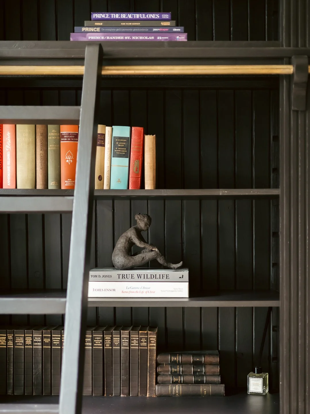 Een zwarte boekenkast met verschillende boeken en een sculptuur van een zittende persoon op een laagte, naast een paar boeken en een fles parfum.
