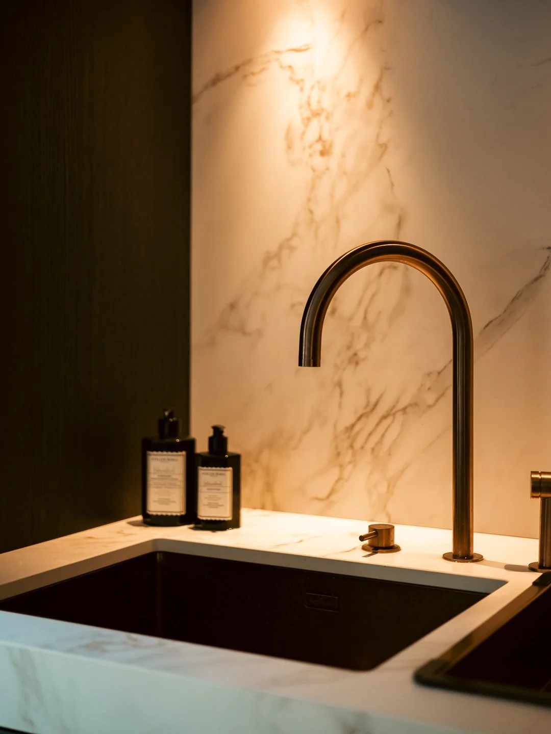 Een zwarte gootsteen met een gouden kran en twee zwarte zeepdispensers op een witte marmeren aanrecht tegen een wit-marmeren muur, binnen een badkamer of keuken.