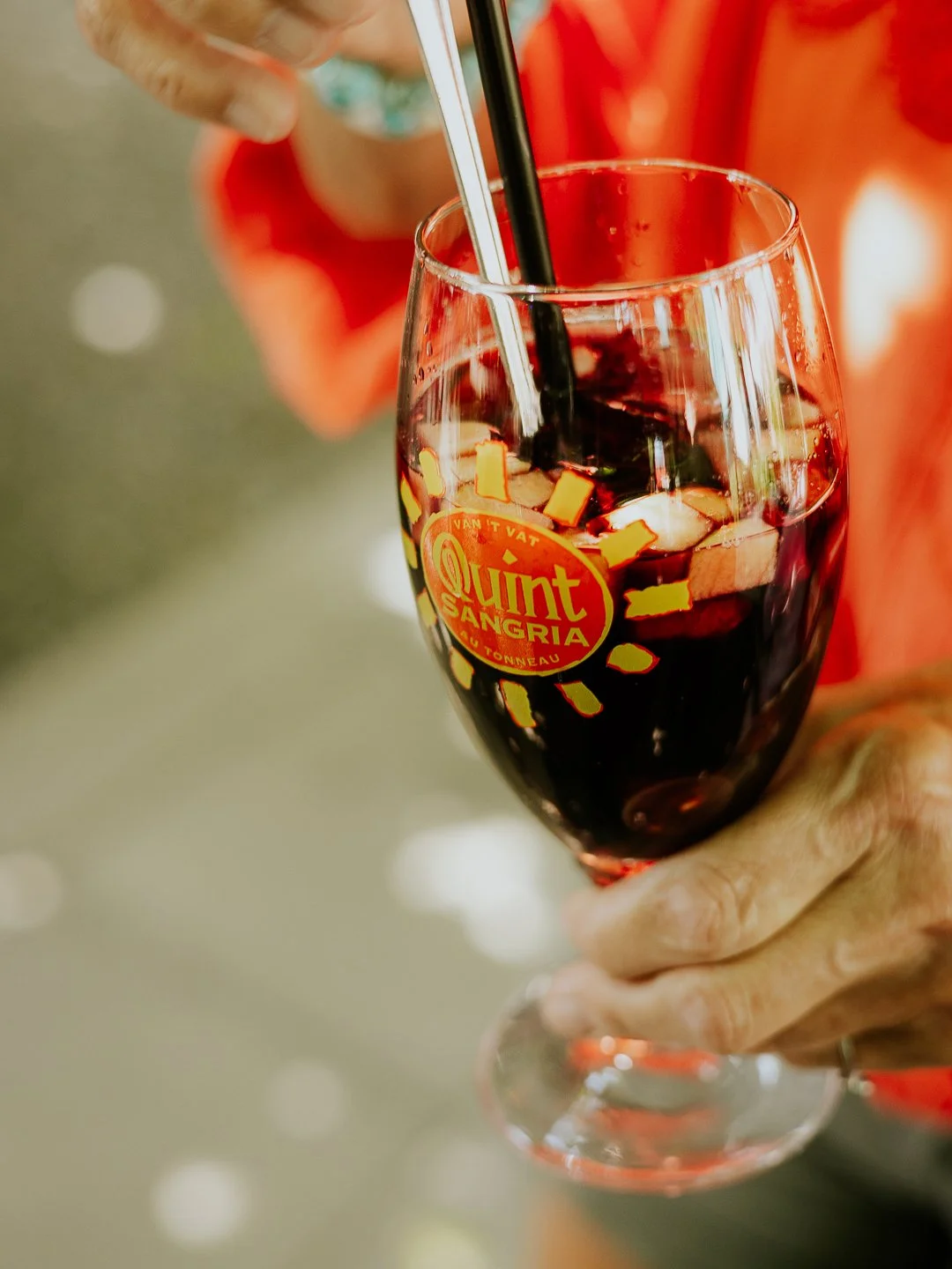 Een hand houdt een glas met een zwarte vloeistof, ijsblokjes en citroenschijfjes, met een logo 'Quint Sangria' op het glas.