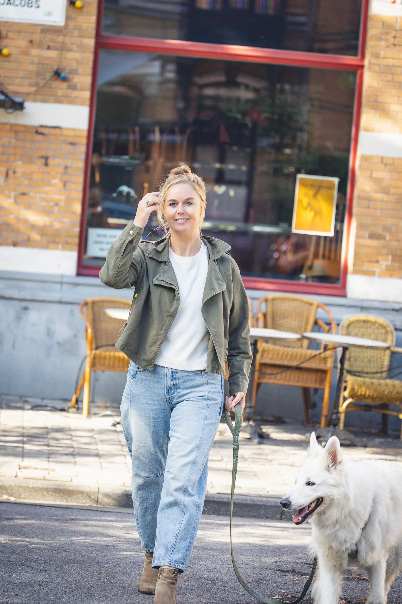 Vrouw die met een witte hond loopt langs een café met buitenzitplaatsen.