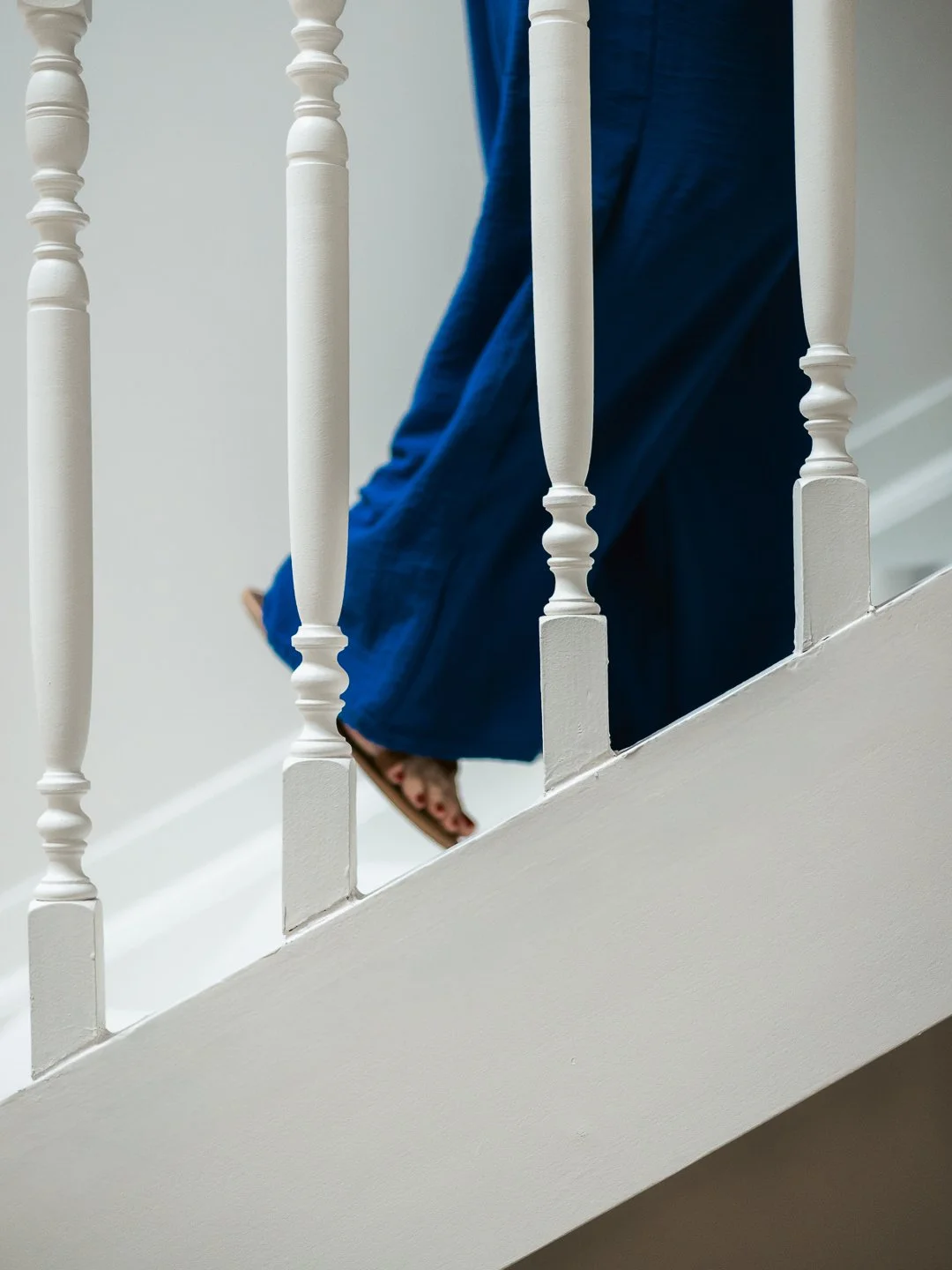 De afbeelding toont een persoon die de trap op loopt, gekleed in een blauwe jurk en slippers. Alleen de onderkant van de persoon is zichtbaar. De trap en leuningen zijn wit.