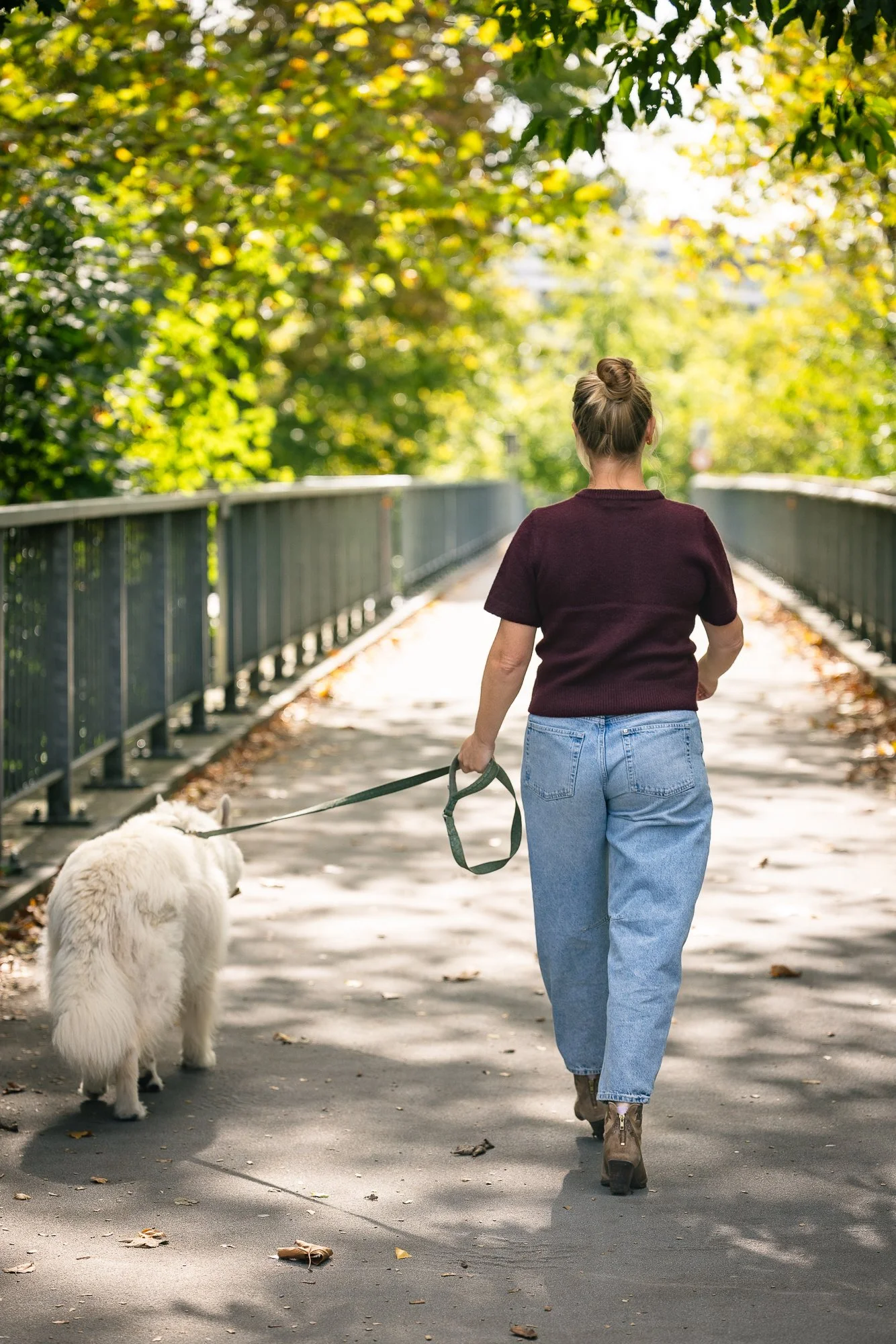Een vrouw wandelt met een witte hond over een schaduwrijke brug met groene bomen aan weerszijden.
