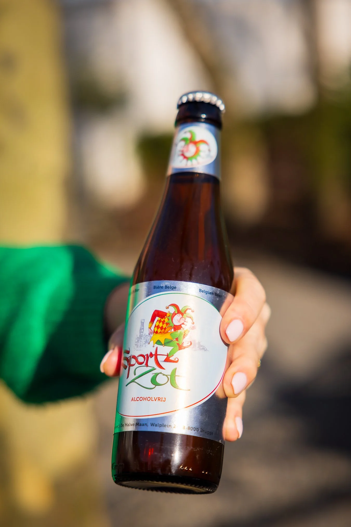 Persoon houdt een fles alcoholvrije Belgisch biertje in de hand. Op het label staat een kleurrijk logo en de tekst 'Sport' en 'ALCOHOLVRIJ'.