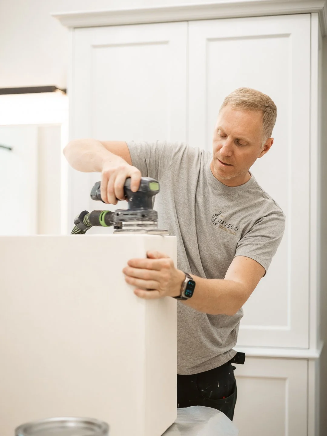 Man werkt met een schuurmachine op een wit keukenmeubel, draagt een grijs T-shirt met het logo 'JAVECO'.