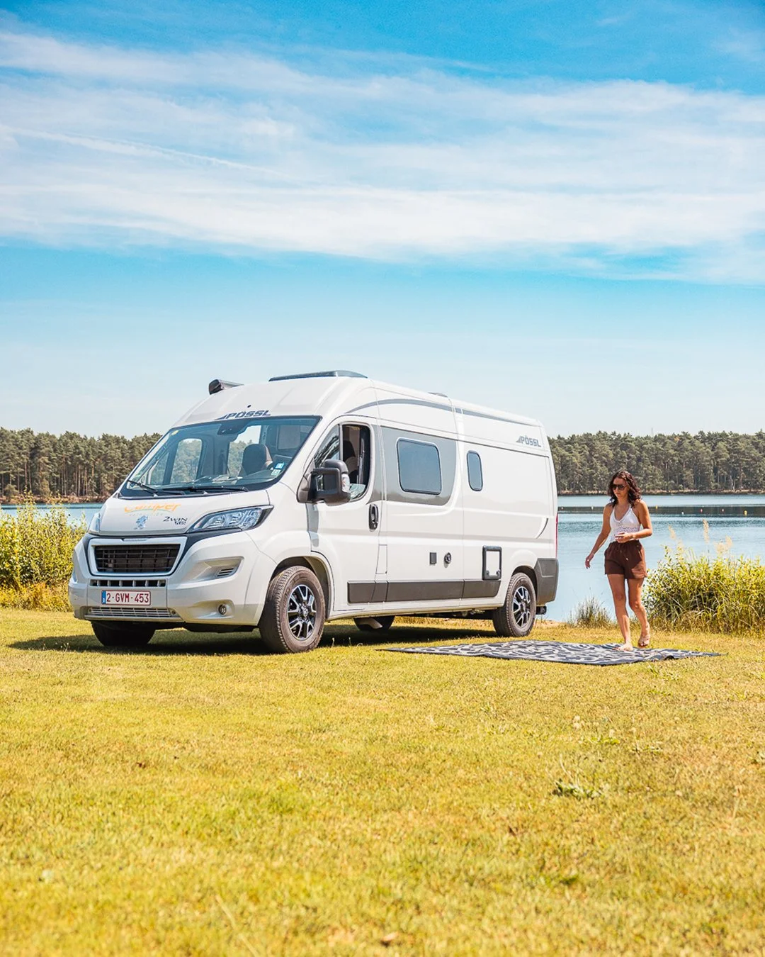 Een witte camper staat op een grasveld bij een meer, met een vrouw in zomerkleding die op een kledendeken wandelt.