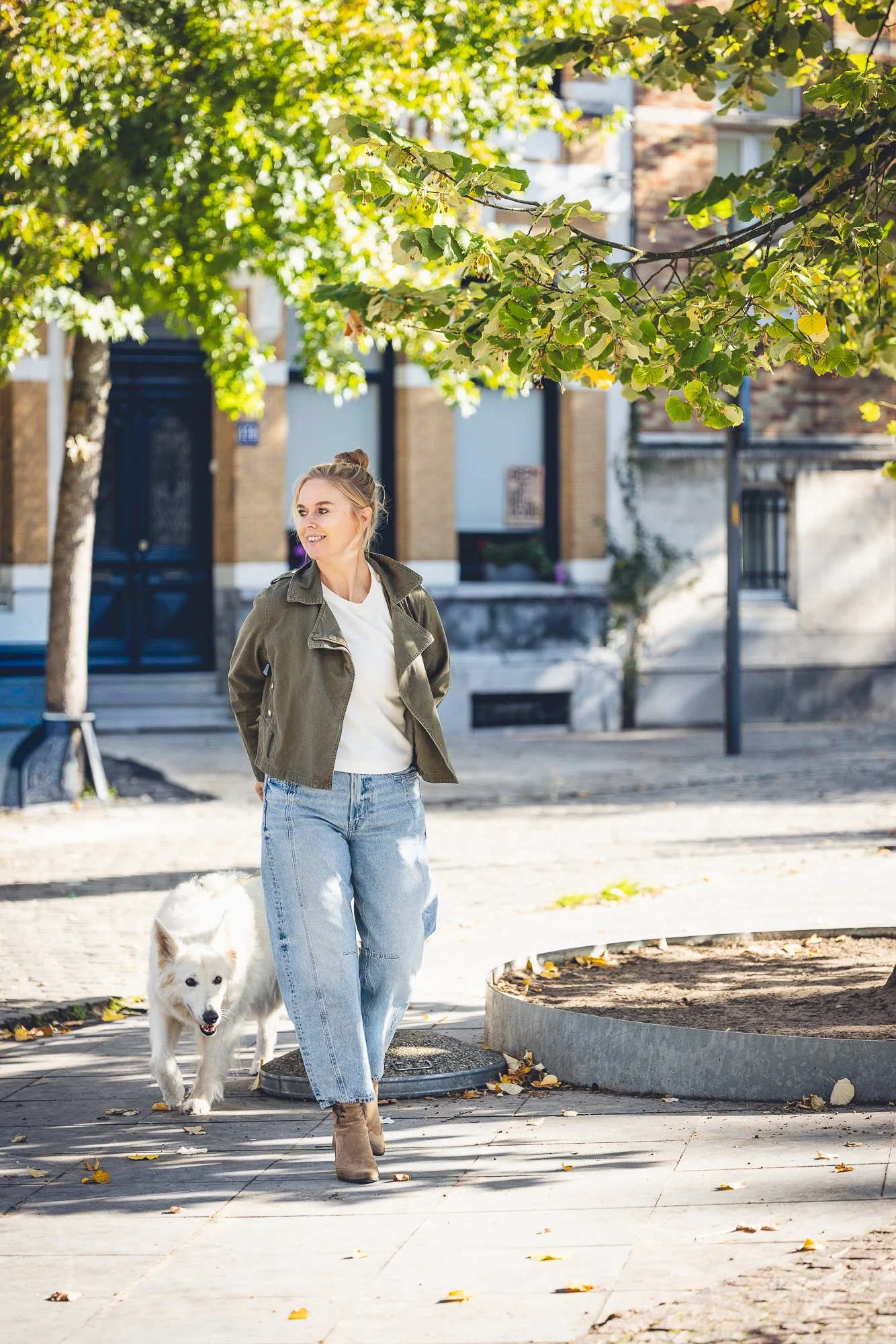 Vrouw die met haar hond wandelt in een park op een zonnige dag, met groene bomen en een stedelijke achtergrond.