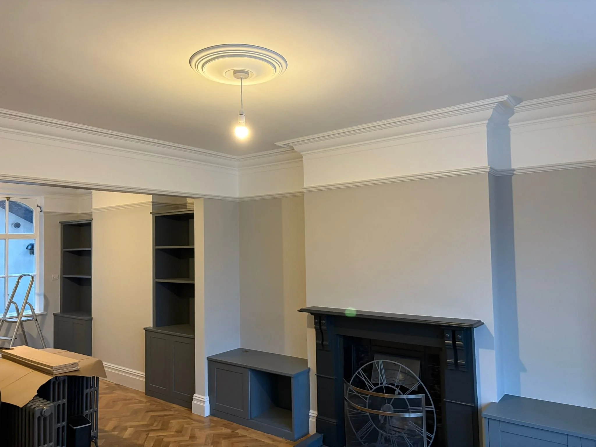 Living room with white walls, decorative crown molding, and a ceiling light fixture. There is a black fireplace with a metal decorative clock in front of it. Built-in gray shelves and cabinets are on the walls, with some furniture and a ladder visibl