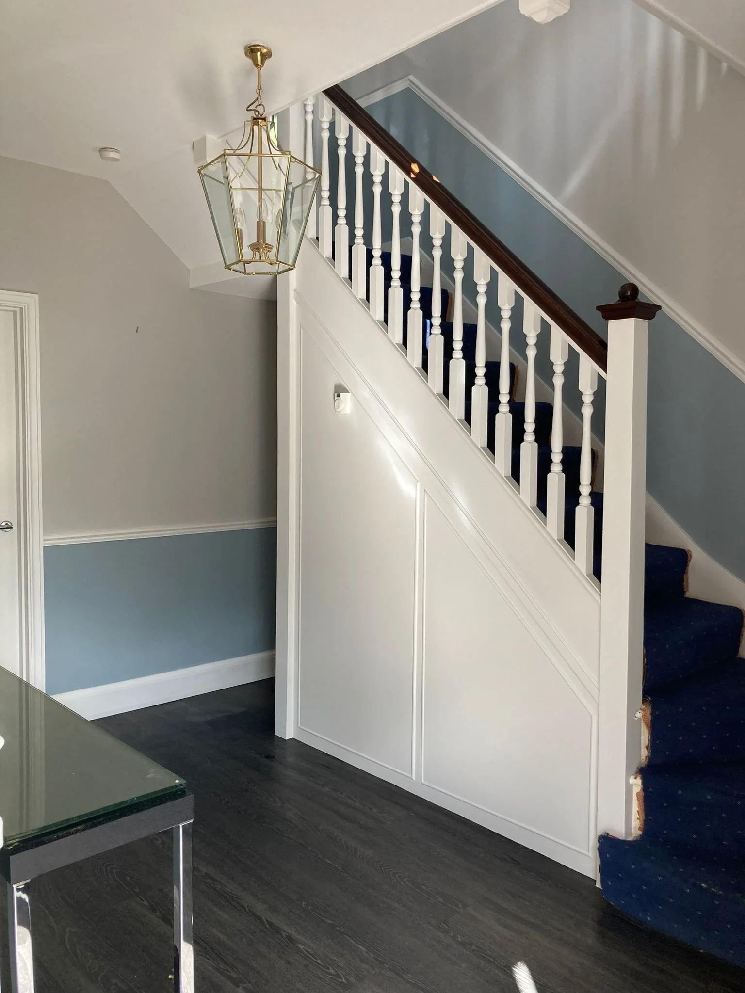 Interior view of a staircase with blue carpet and white balustrade, adjacent to a white wall with blue paneling, and a black wood floor. A gold hanging lantern-style light fixture is above.
