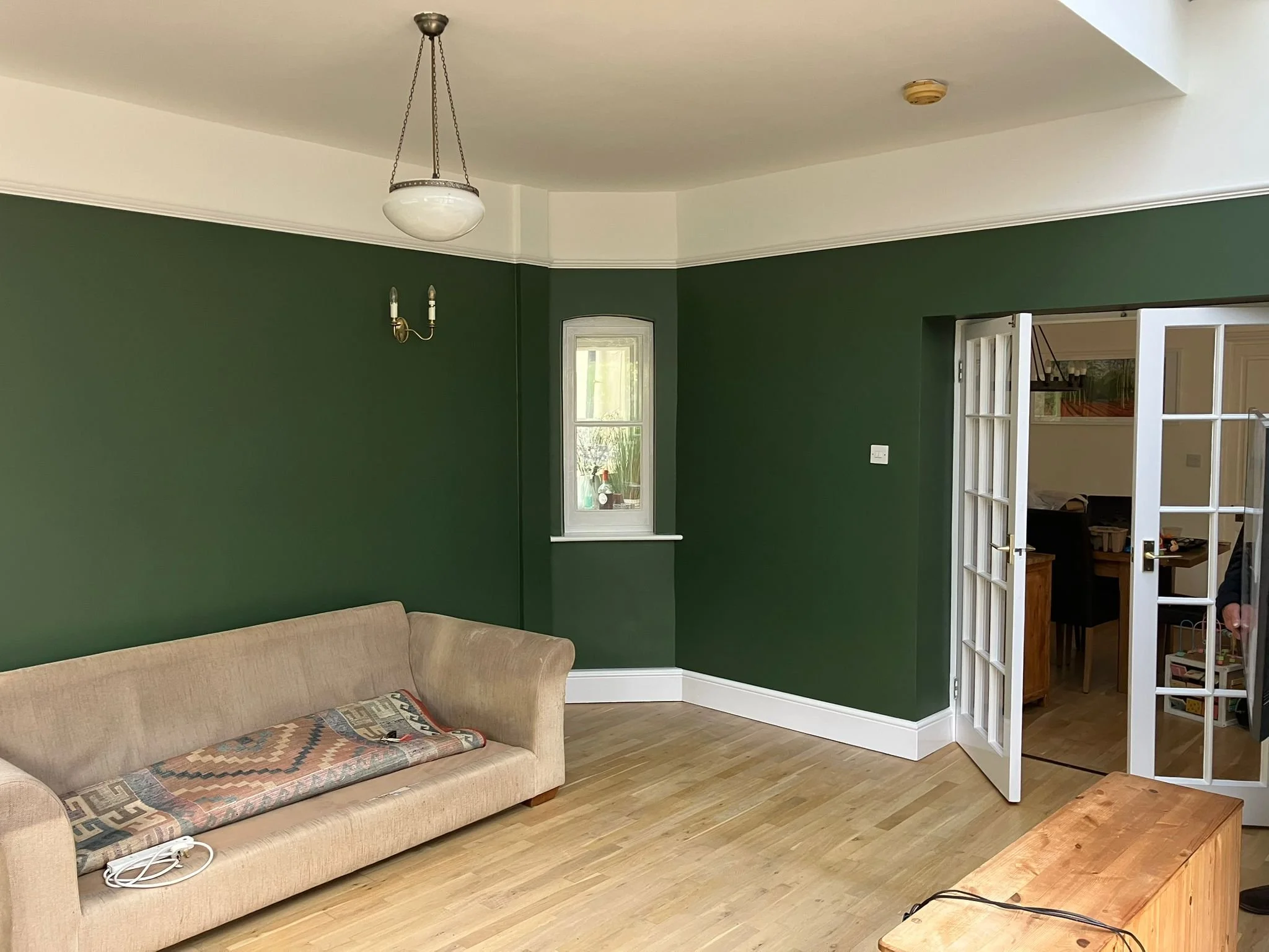 Living room with green walls, beige sofa, wooden floor, window, and open French doors leading to a dining area.