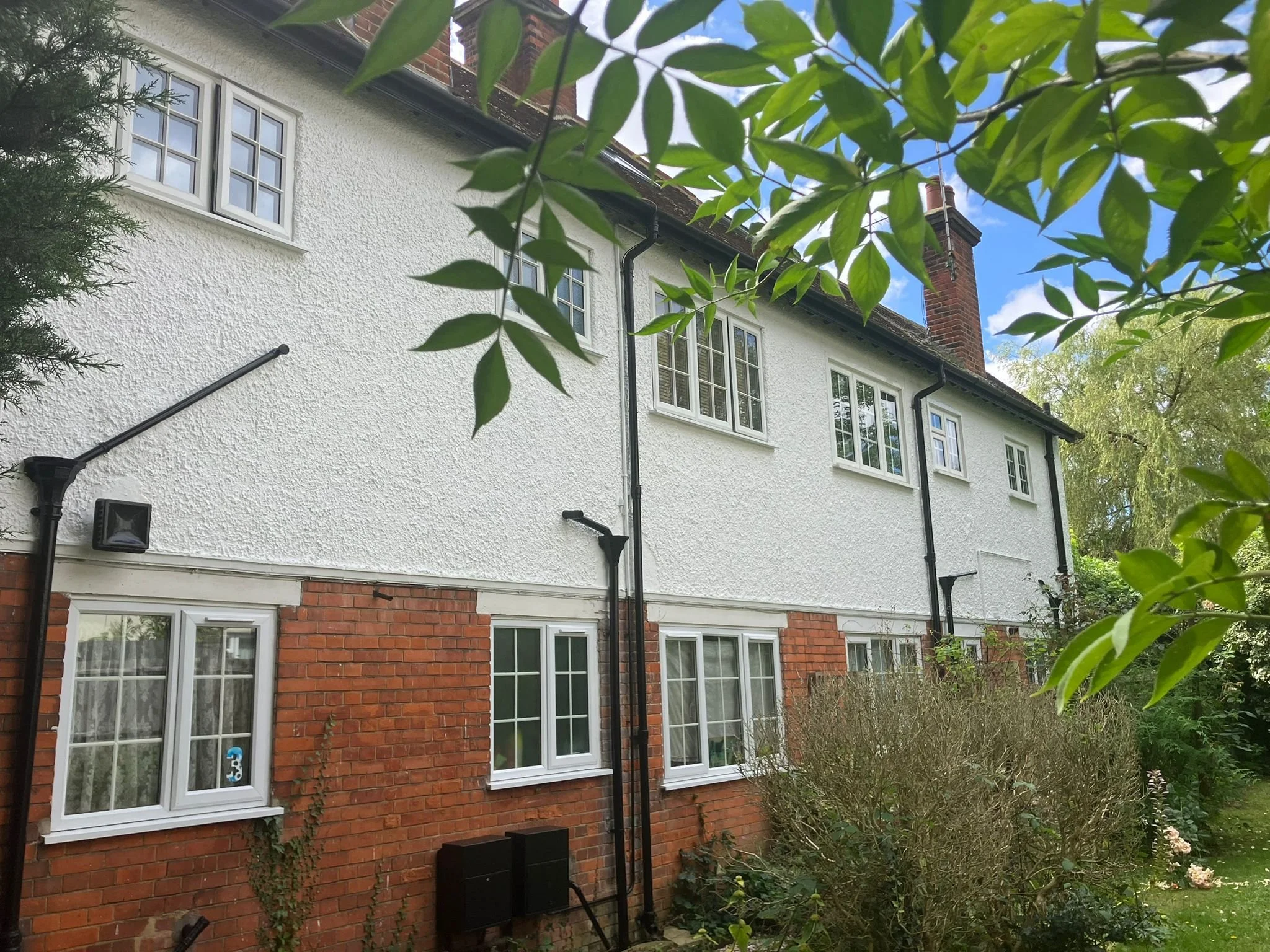 Two-story house with white textured upper walls, red brick lower walls, multiple white-framed windows, black gutters and downspouts, and a chimney. Green leaves and trees are visible in the foreground and background.