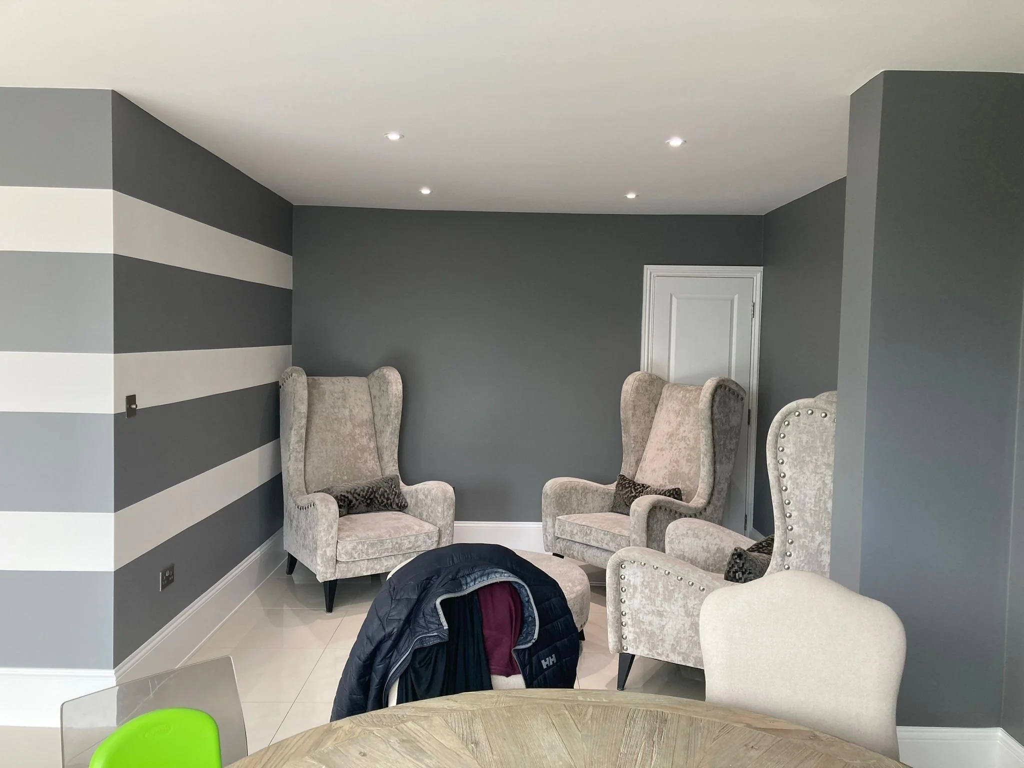 Living room with gray and white walls, four beige armchairs, a white door, and a round wooden table with a jacket hanging on a chair in the foreground.