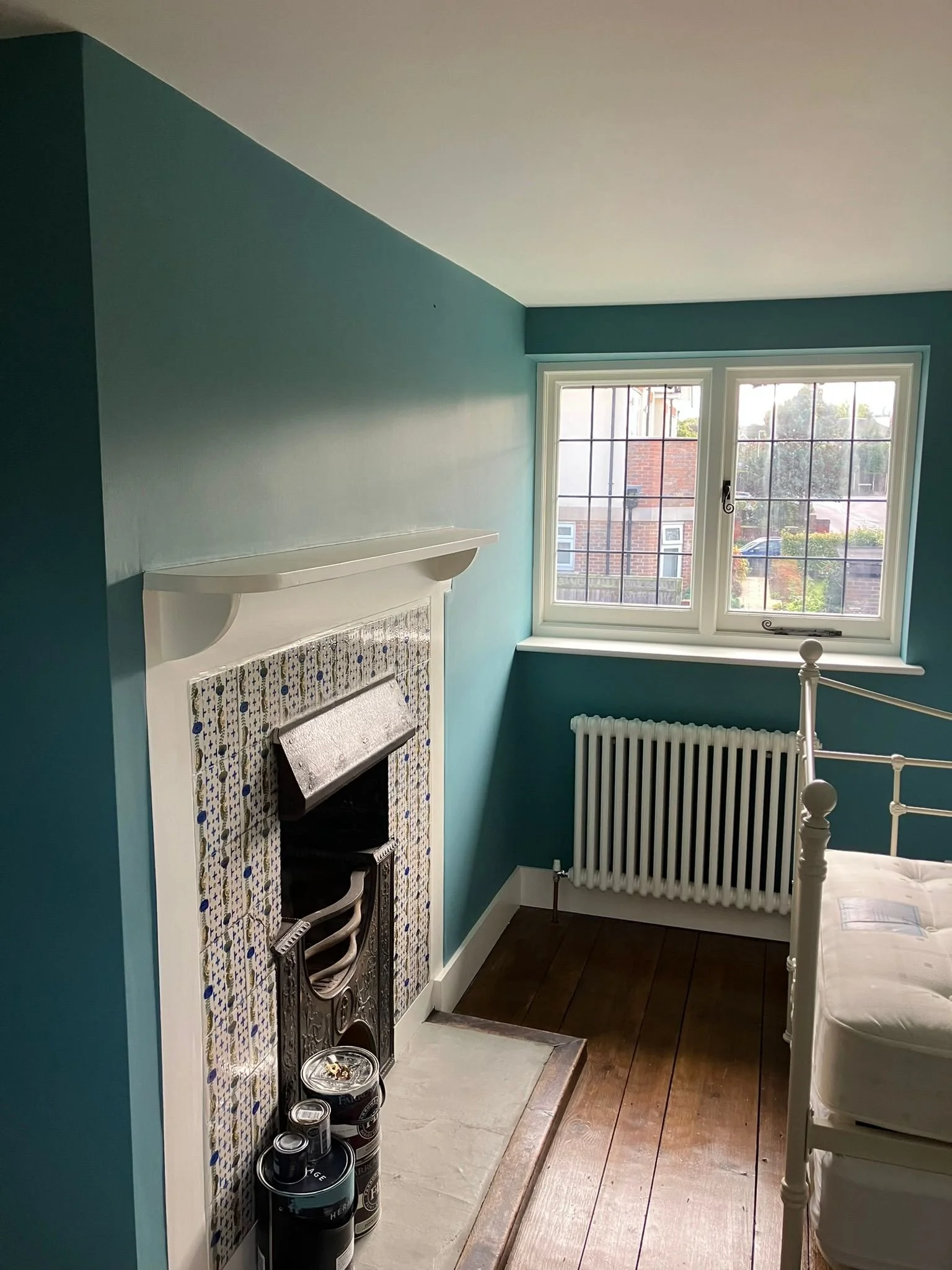 A corner of a bedroom with teal walls and a white window showing an outdoor scene, a white radiator below the window, a section of a white bed frame, and a fireplace with decorative tile surround and cans of paint nearby.