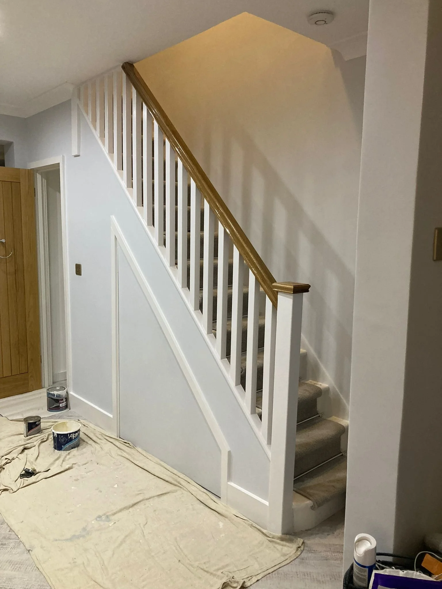 Indoor staircase with wooden handrail and white spindles, tan carpeted steps, paint cans on a drop cloth, and painting supplies nearby.