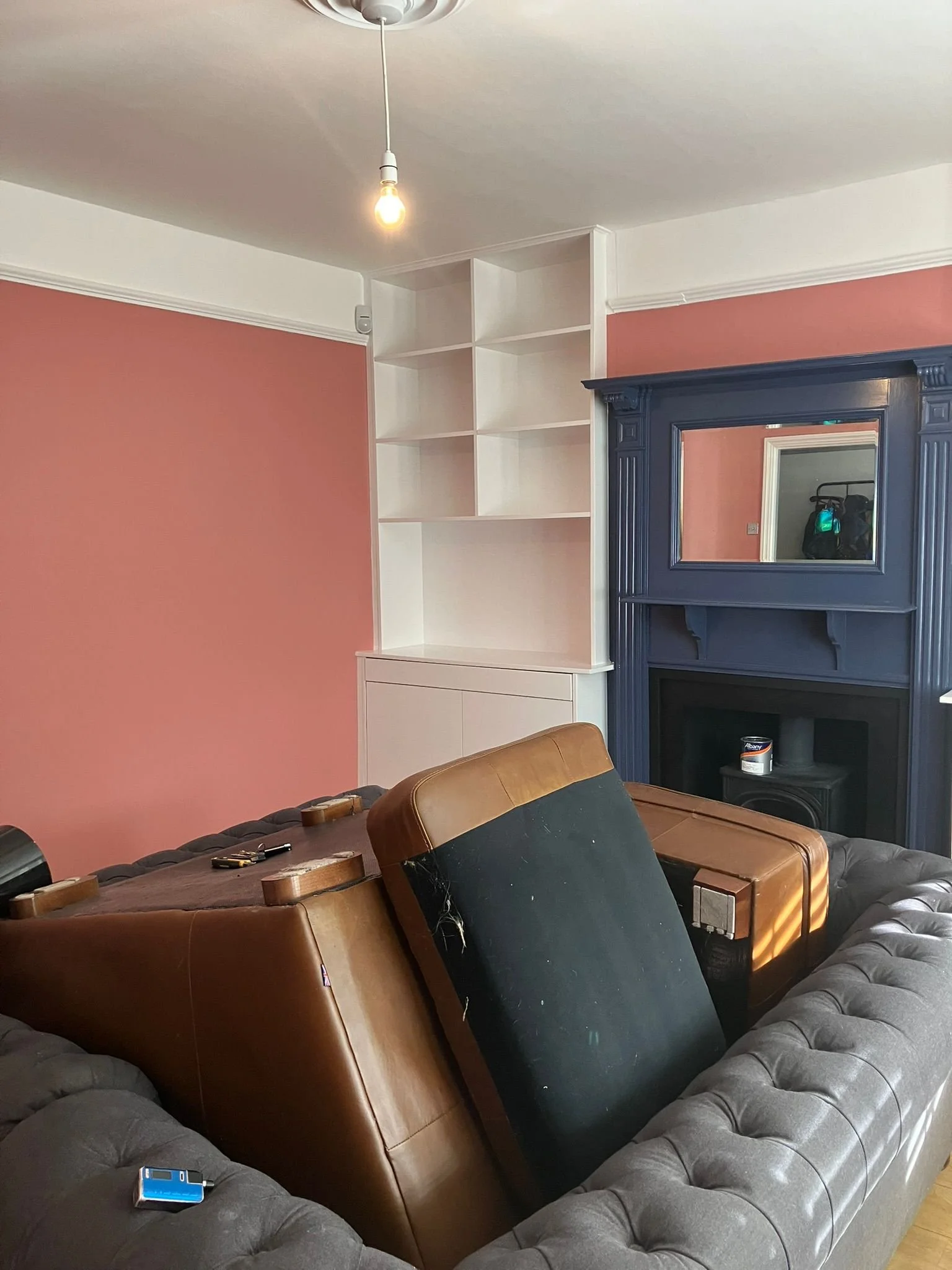 Living room with a pink wall, a white corner shelving unit, a black fireplace with a wood mantel and mirror, a gray tufted sofa, and two brown chairs upside down on the sofa.