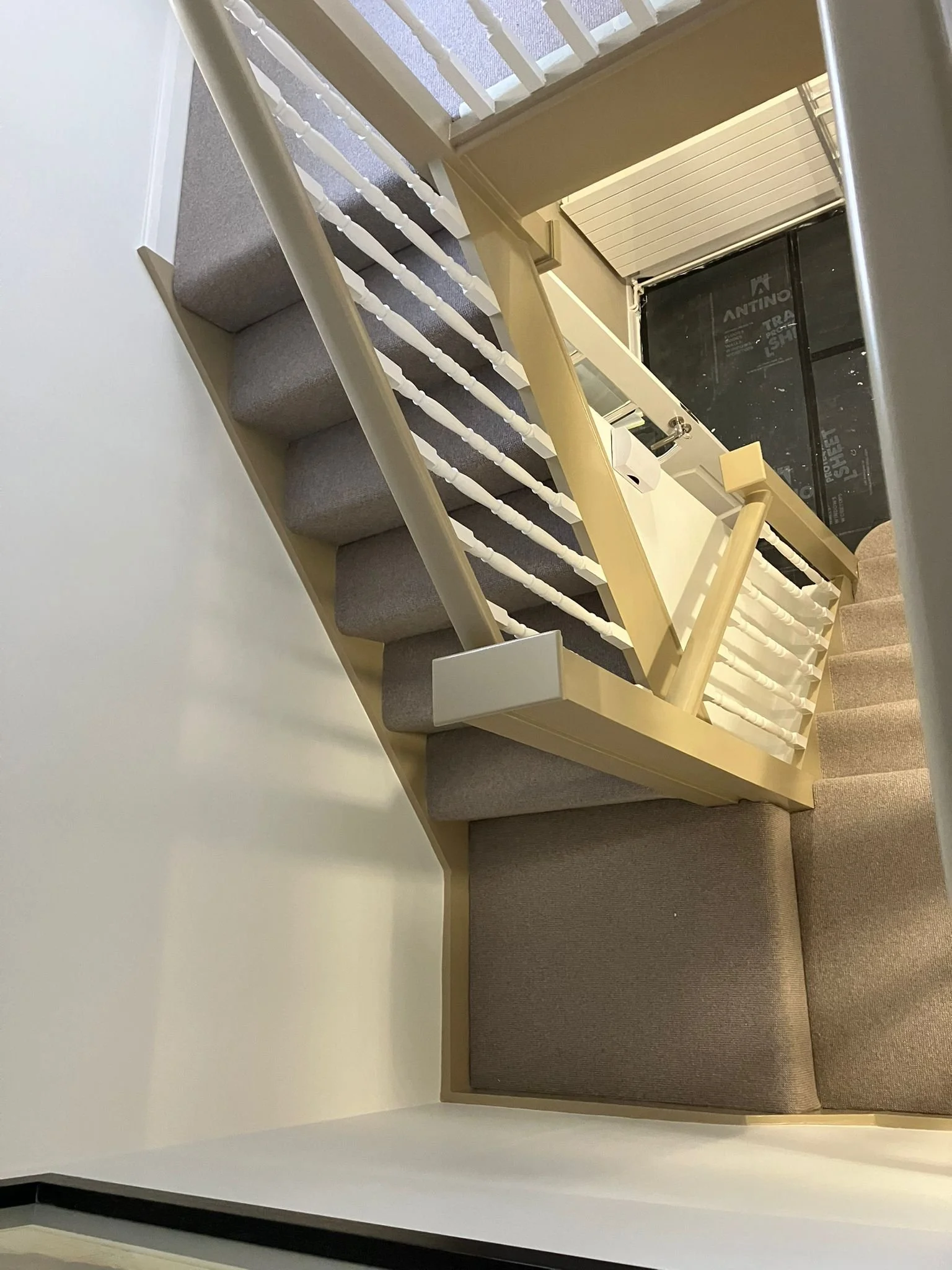View of a staircase with beige painted railings, grey carpeted steps, and white spindles, leading to a corridor with a white coat of paint, and in the background, a black construction barrier at a building site.