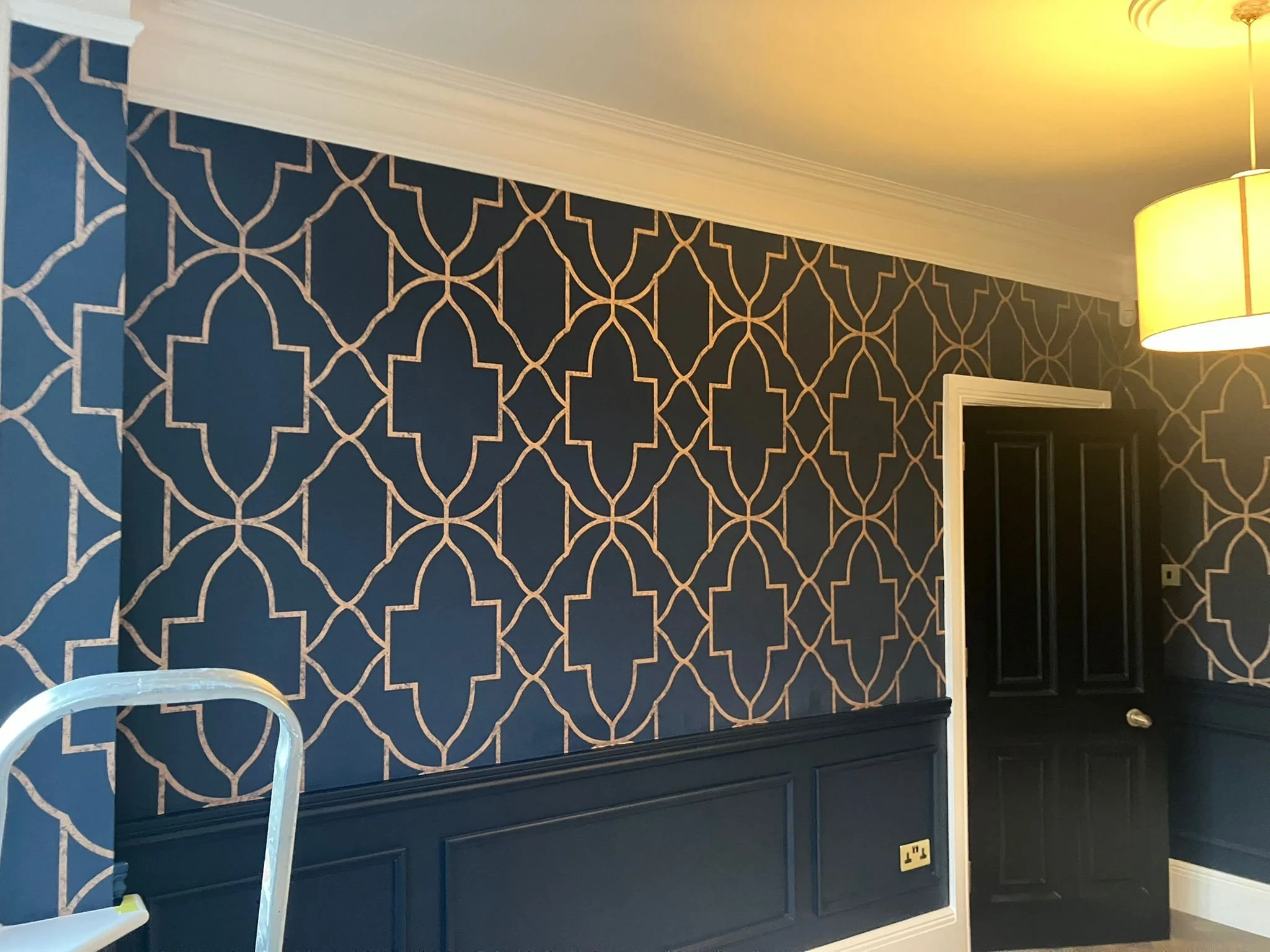 Interior of a room with navy blue wallpaper featuring gold geometric lattice pattern, black door, white trim, and a ceiling light with a yellow lampshade.