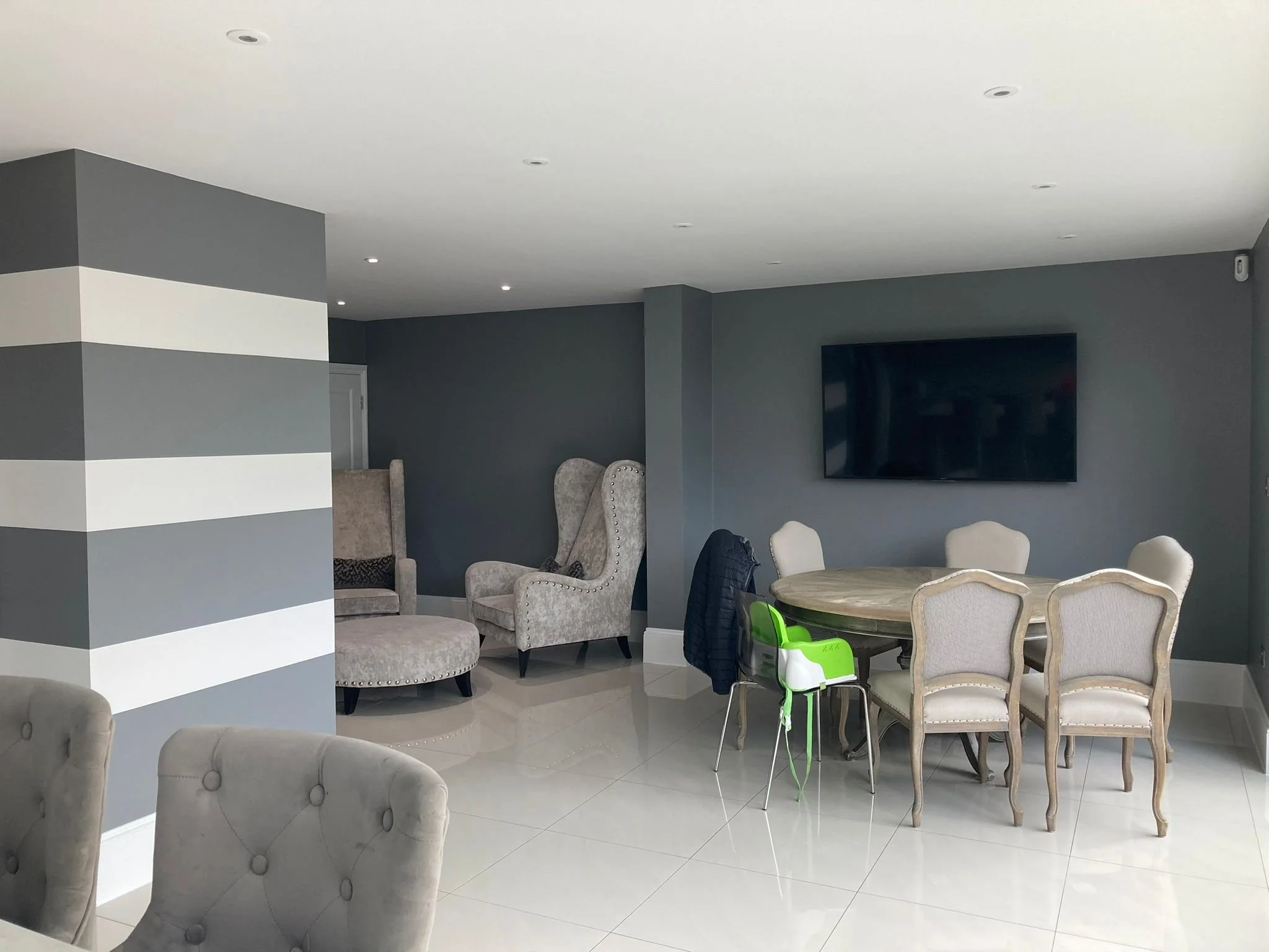 Living room with gray walls, a TV mounted on the wall, a round dining table with six chairs, and gray upholstered armchairs and high-back wing chairs in the background.