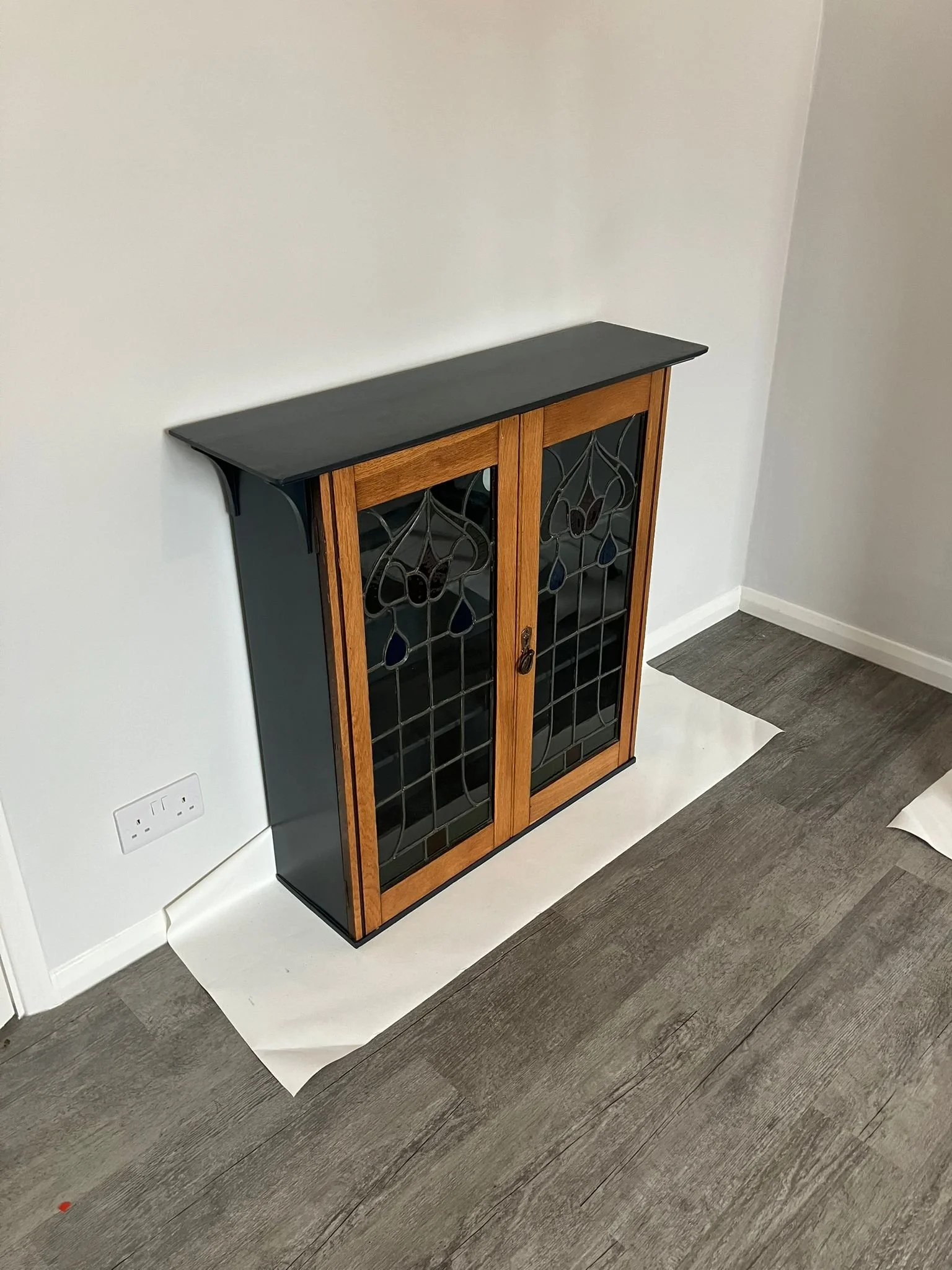 A wooden cabinet with glass-paneled doors featuring decorative stained glass designs, topped with a black shelf, placed against a plain white wall on a hardwood floor.