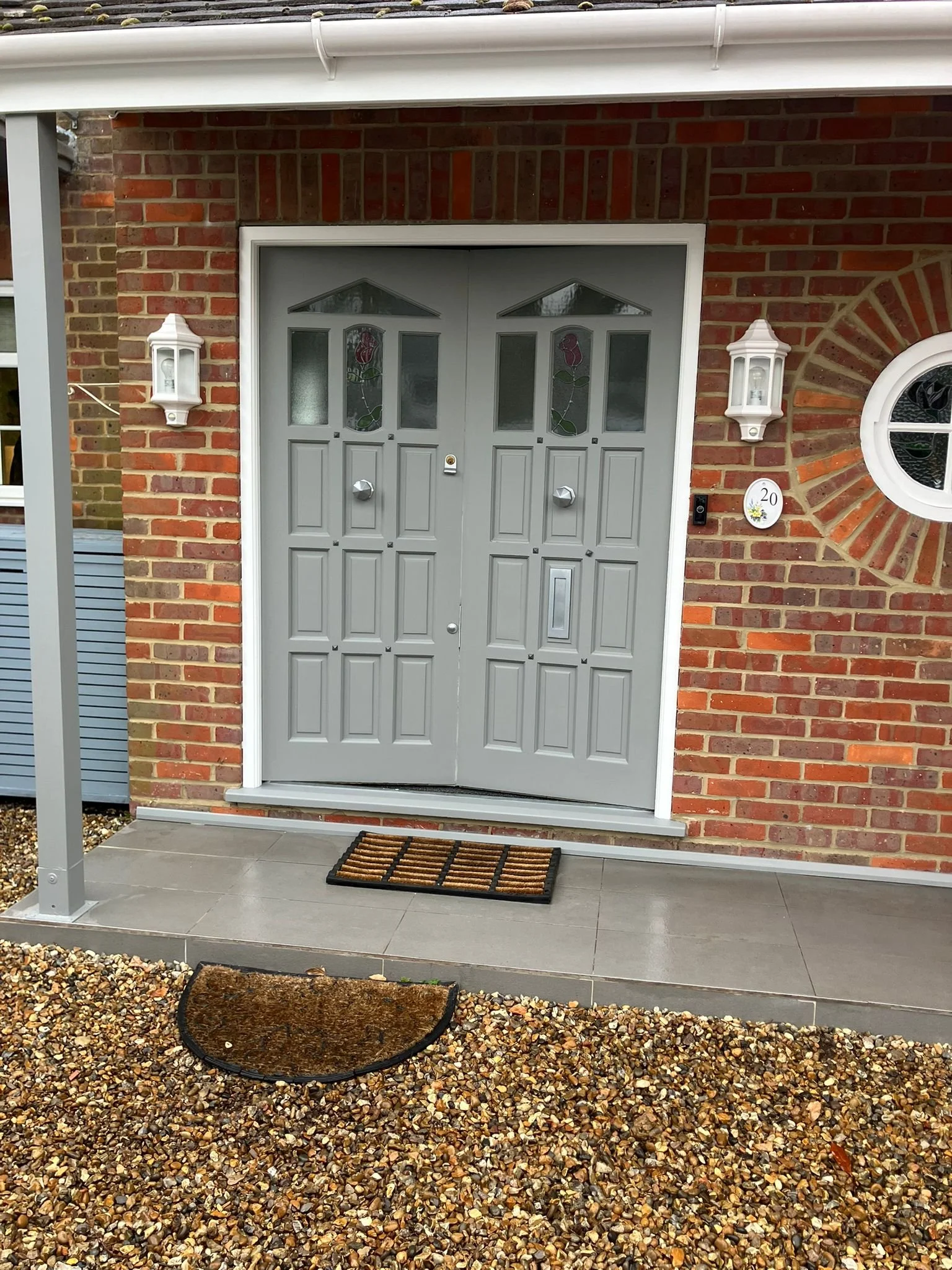 Front entrance of a brick house with a gray door, two lantern-style wall lights, a doormat, and a pebble pathway.