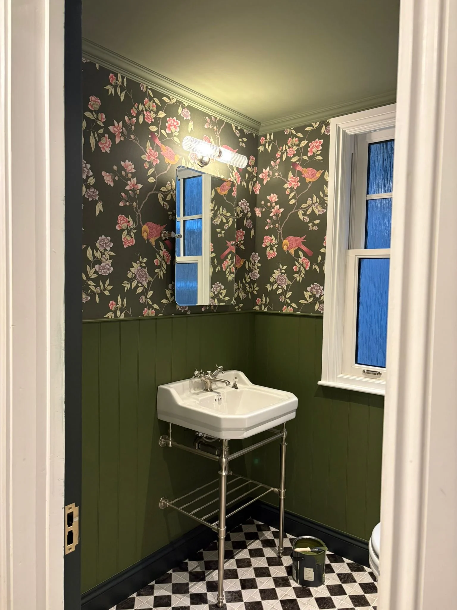 Small vintage bathroom with floral wallpaper, a green wainscot, a wall-mounted mirror, and a white sink with exposed metal piping, next to a window with frosted glass.