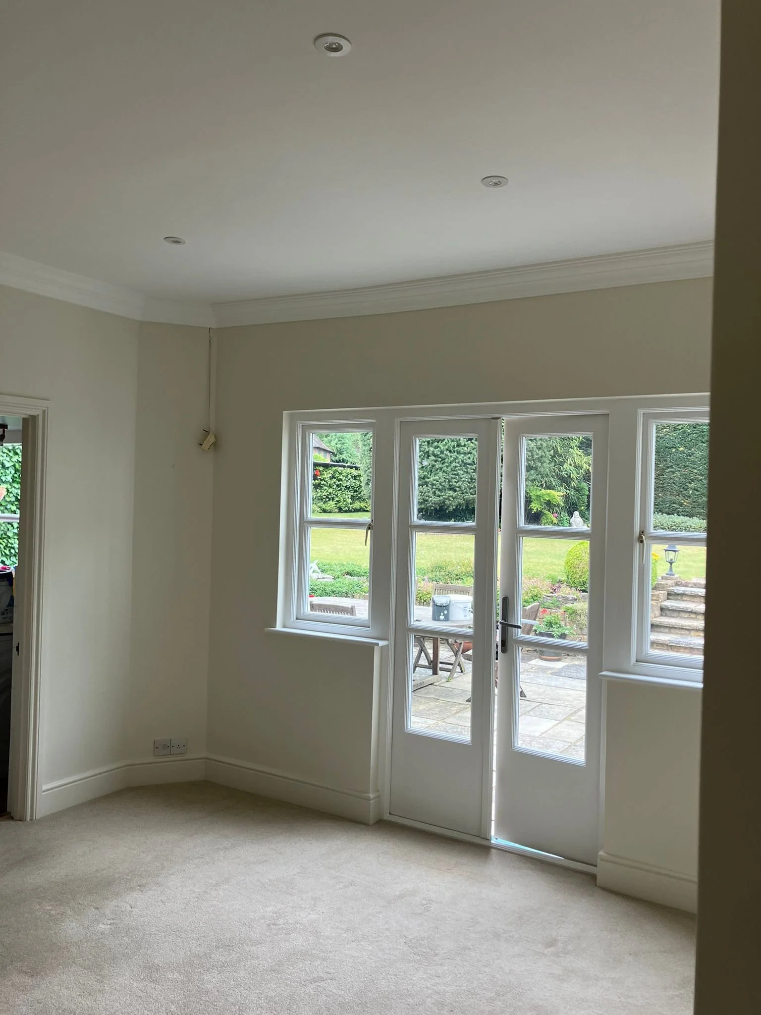 Empty room with white walls, beige carpet, large windows, and a glass door leading to an outdoor patio with garden furniture.