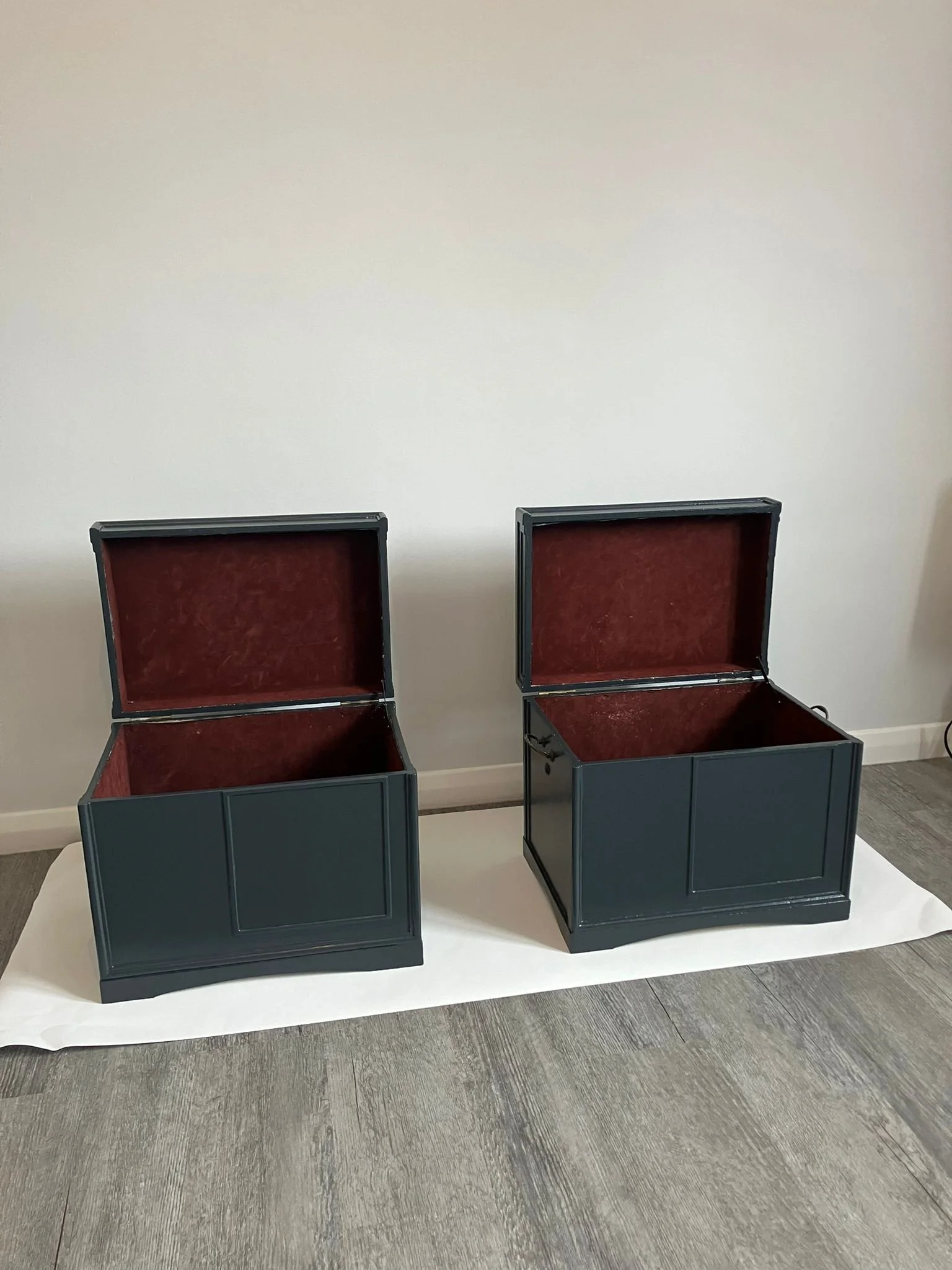 Two empty blue display cases with open lids and red velvet interiors, placed on a white sheet on a wood floor against a plain white wall.