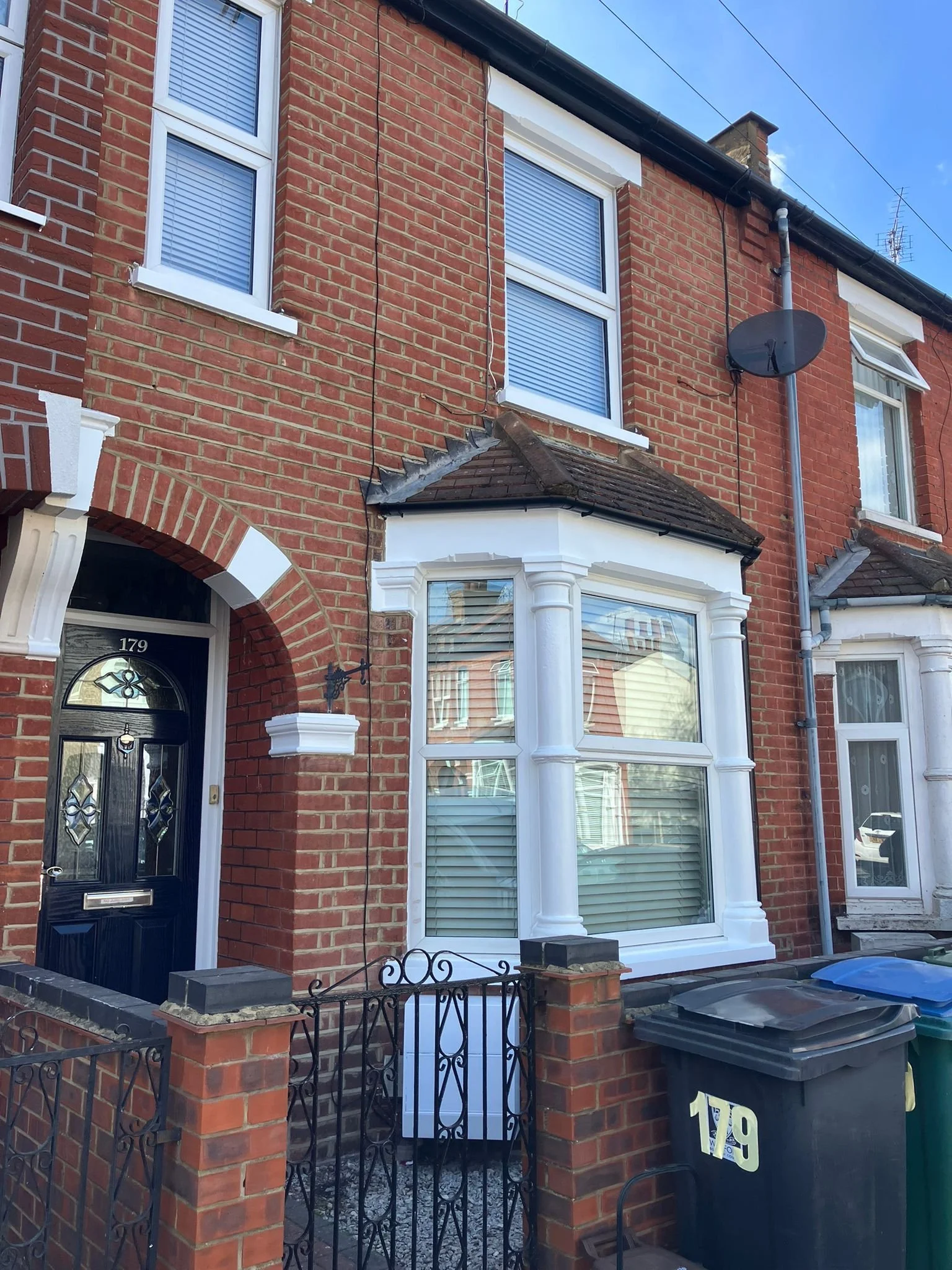 A brick townhouse with white decorative trim around a bay window and a black front door, numbered 179, with a small black iron gate and brick fence in front, alongside trash bins.