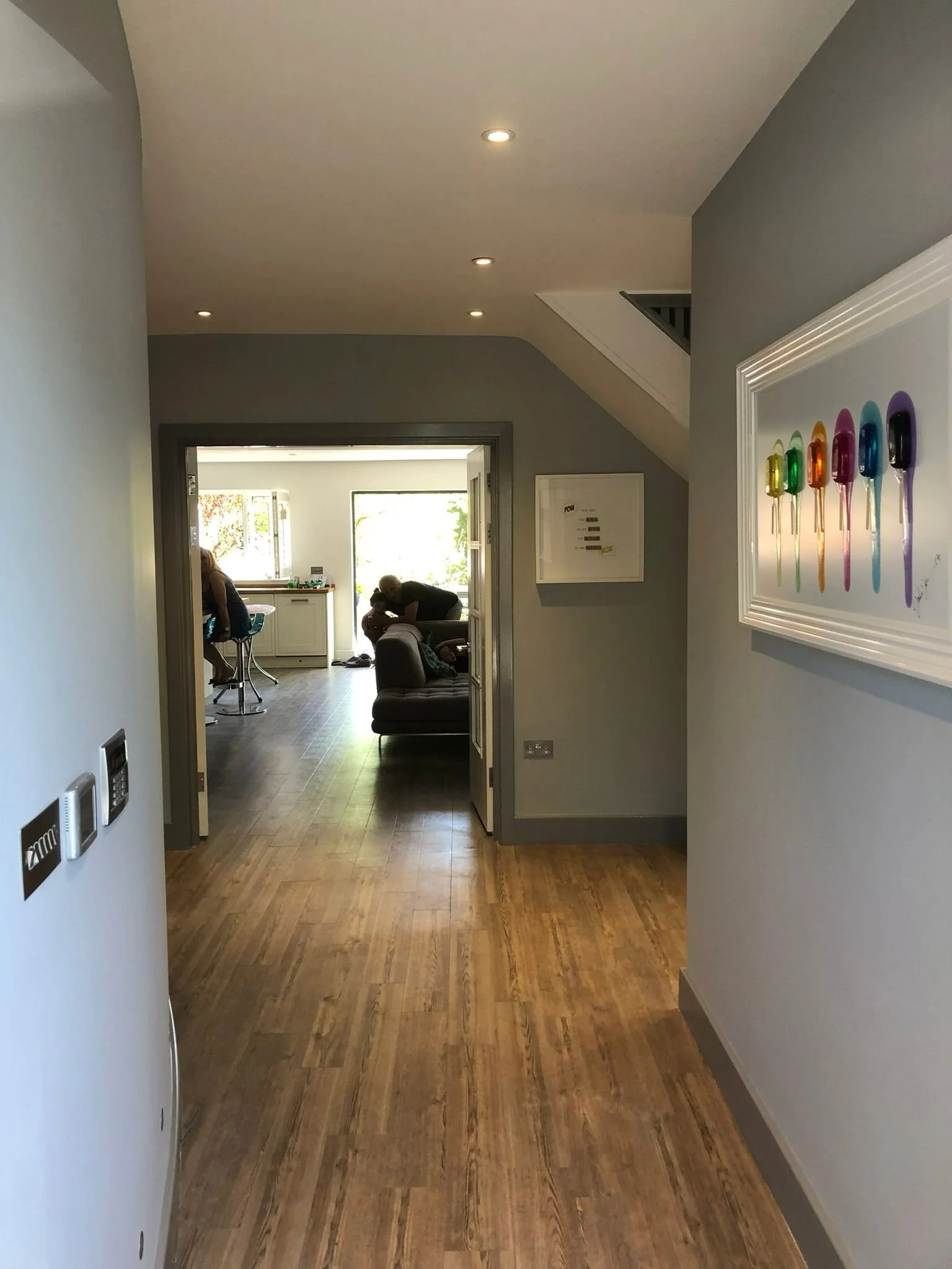 View of a hallway leading into a living room with people sitting and talking, natural light coming through the windows, colorful ice cream art on the wall, wooden flooring, and gray walls.