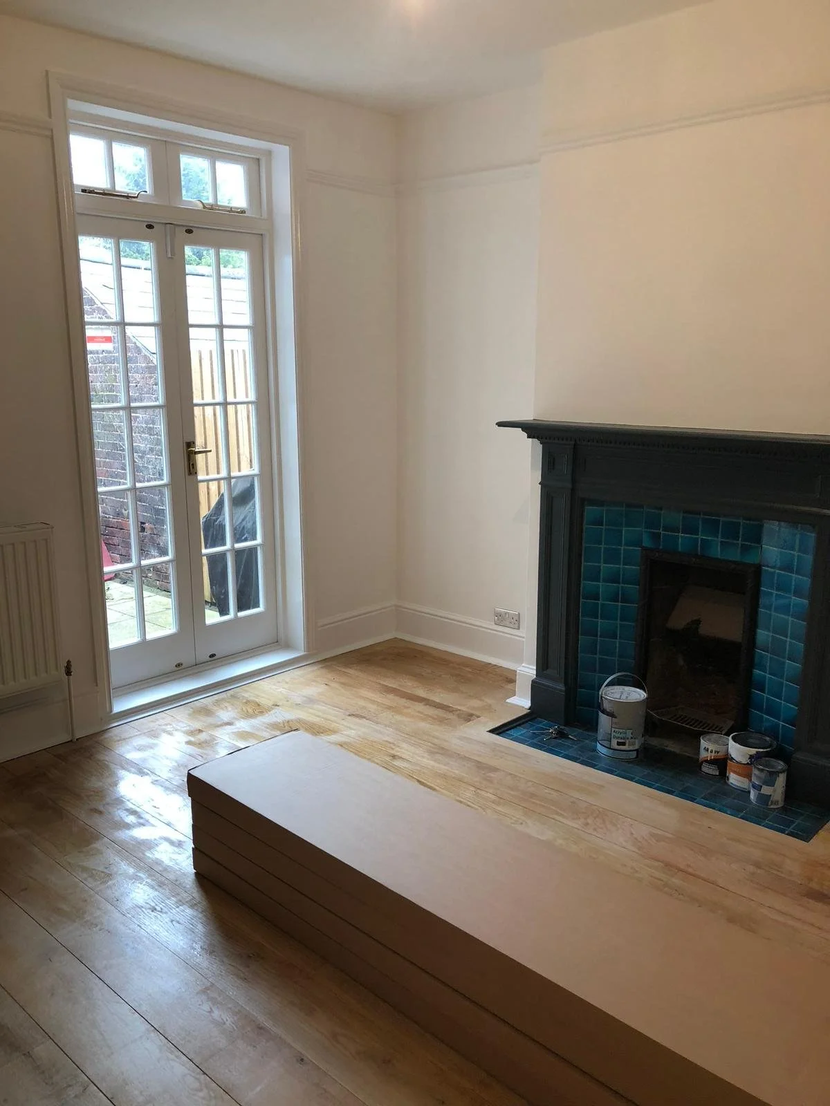 Empty living room with a fireplace with blue tiles, a stack of wooden planks on the floor, and a glass door leading outside.