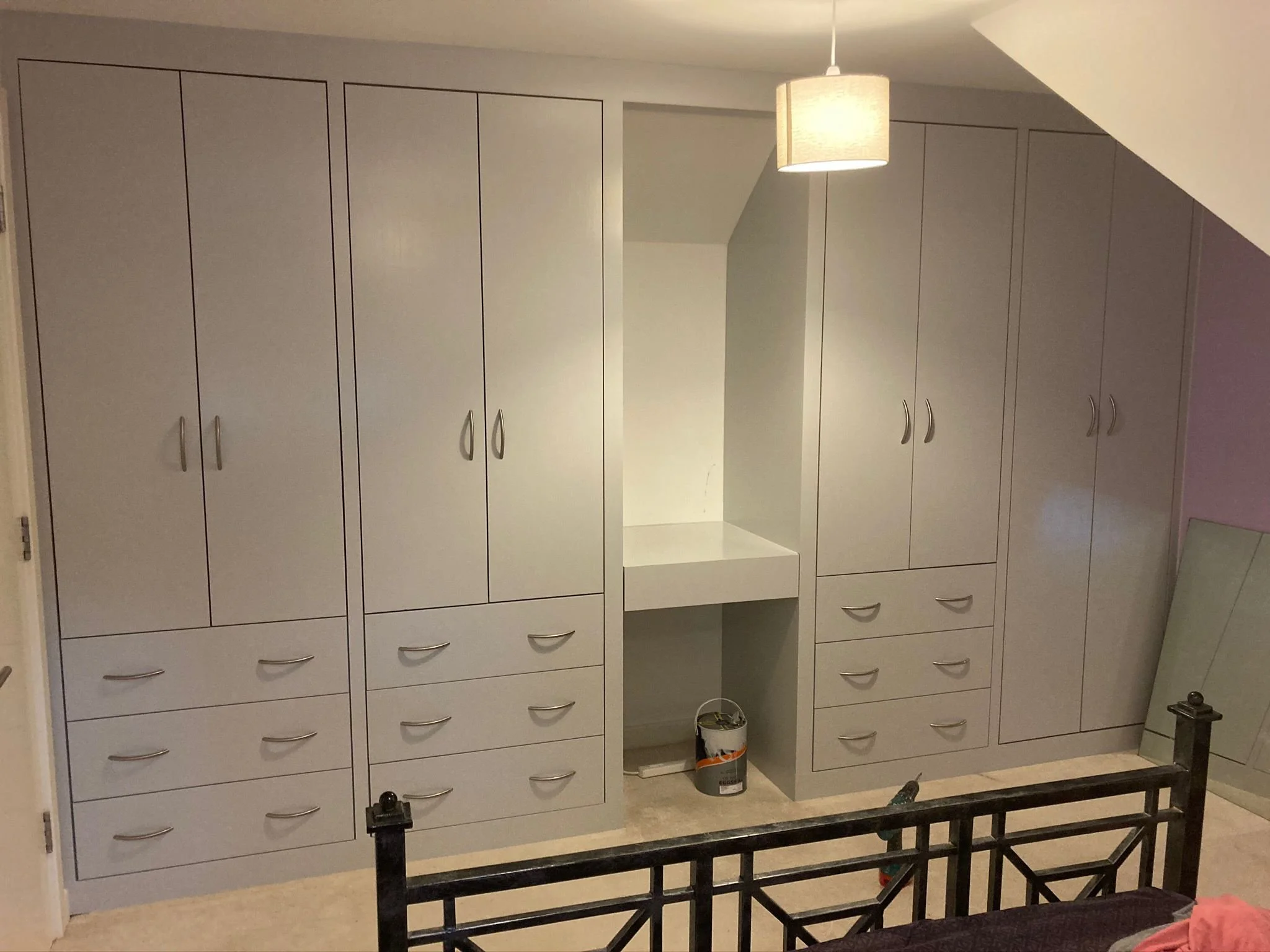A bedroom closet with built-in cabinets and drawers, with a small open shelf in the middle. There is a light fixture hanging from the ceiling, and a bucket on the floor below the shelf. A mirror is leaning against the wall on the right side.