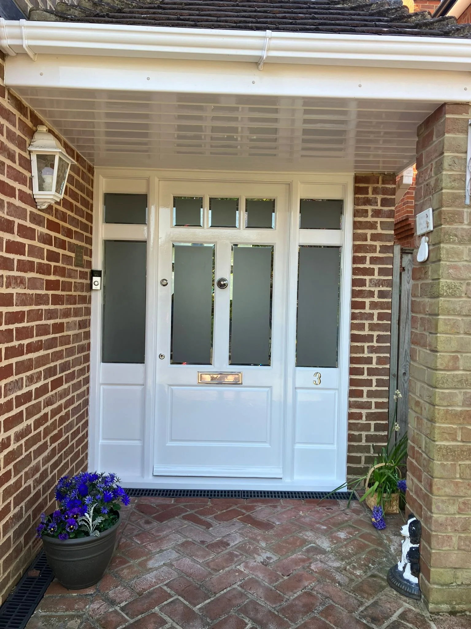 White front door with frosted glass panels, flanked by brick walls and decorated with potted flowers and garden ornaments.