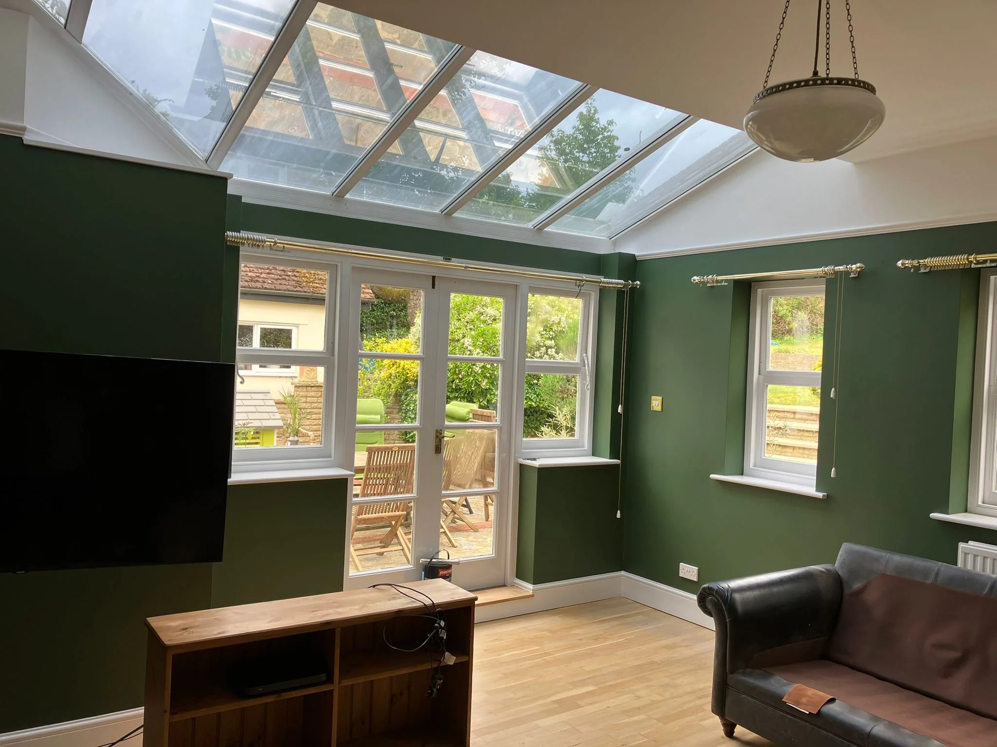 Living room with green walls and wooden flooring, large glass doors leading to a patio, and a skylight ceiling. Furniture includes a wall-mounted TV, a wooden cabinet, and a black leather sofa.