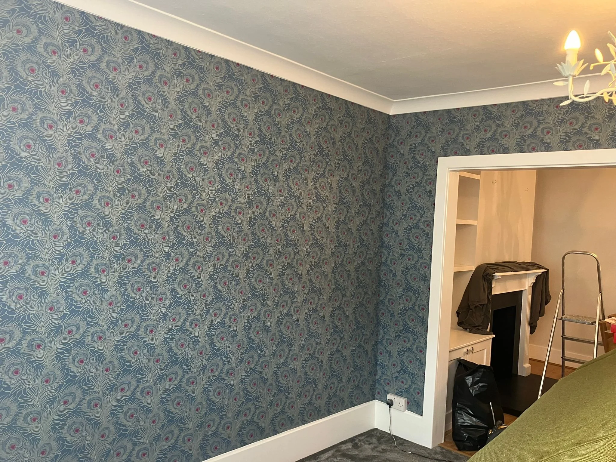 A room with blue patterned wallpaper with pink accents, a white ceiling with crown molding, a hanging light fixture, and a doorway leading to another room with a small fireplace, shelving, a stepladder, and various items.