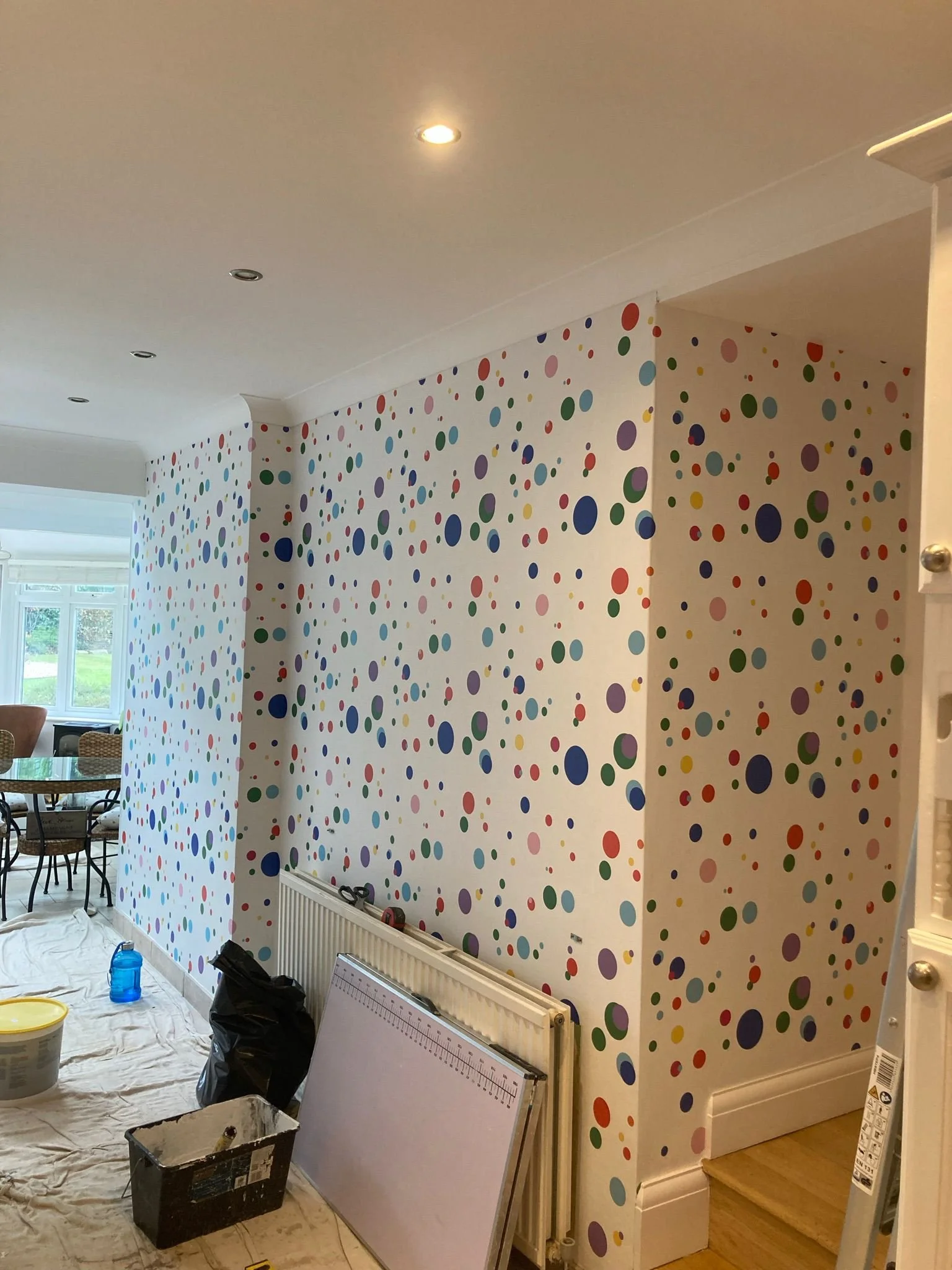 Room with polka dot wallpaper in various colors, some furniture, paint supplies, and a view of a dining area with windows.
